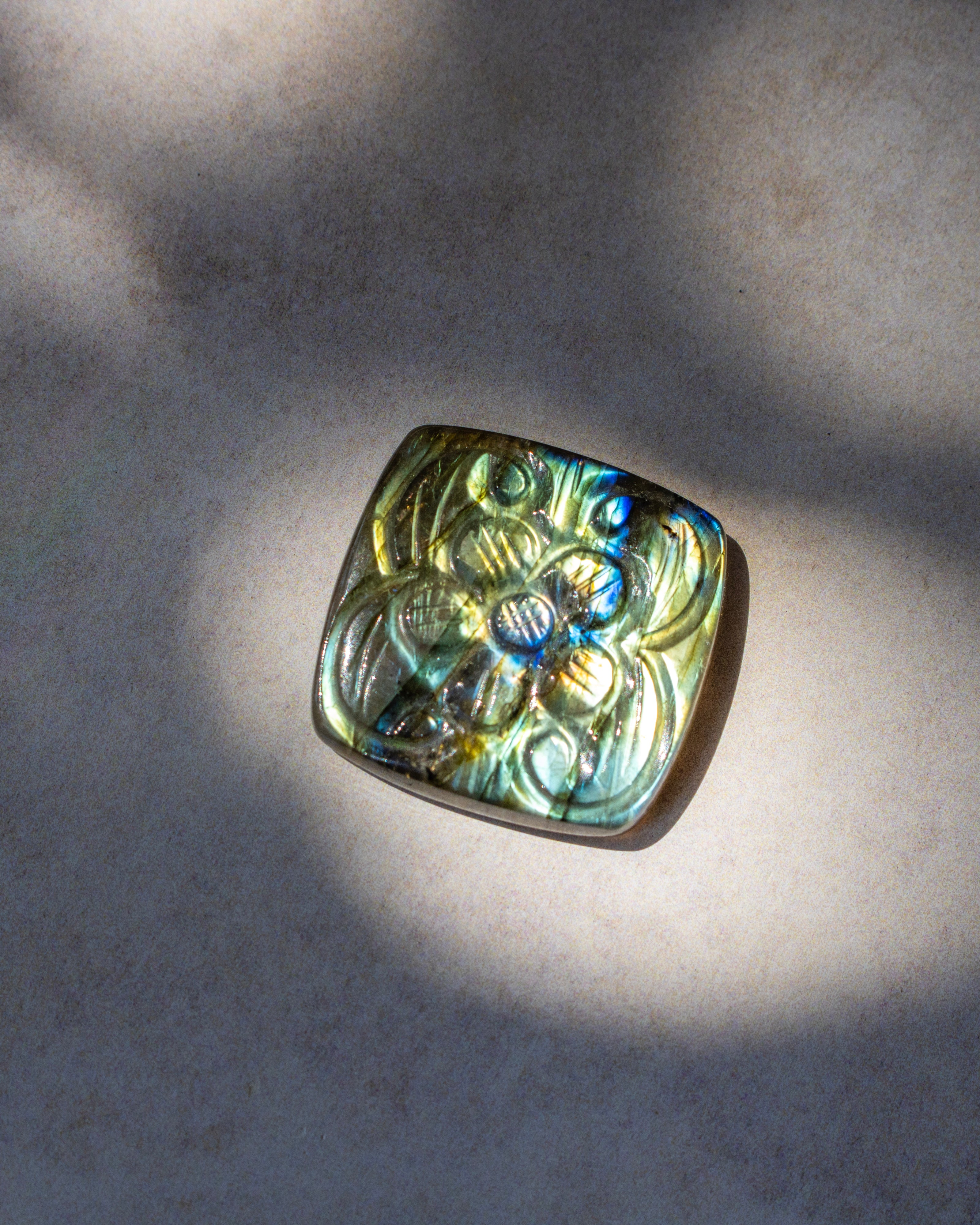 A square pendant with rounded corners featuring an embossed floral pattern, made of iridescent labradorite stone that shifts between green, blue, and gold, resting on a textured beige surface under dappled sunlight.