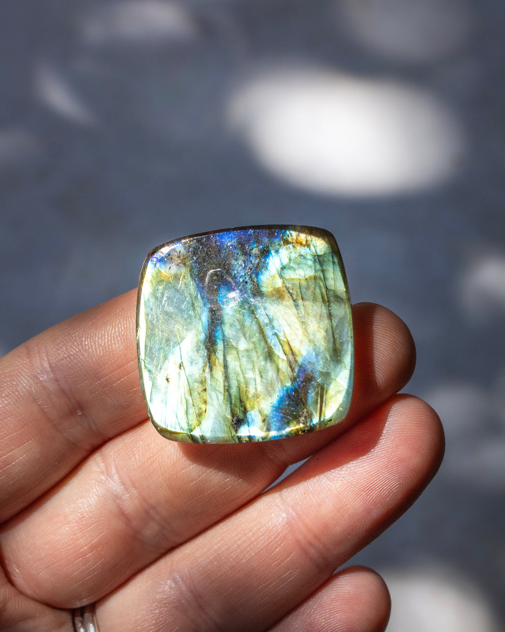 A polished square labradorite gemstone with iridescent blue, green, and gold flashes, held in a person's hand against a blurred gray background with dappled light.