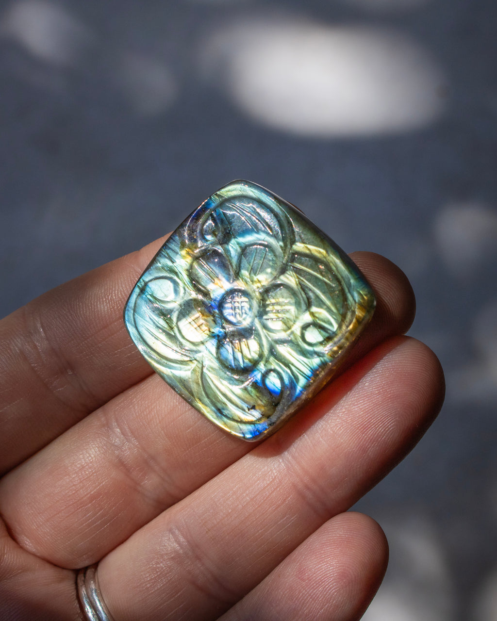 A hand holding a square iridescent glass bead with a raised floral pattern, showing shimmering blue, green, and gold tones against a blurred gray background.