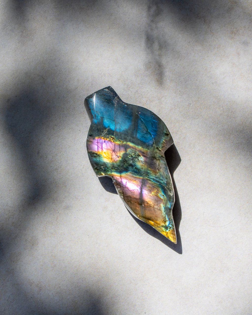 A polished labradorite stone pendant with a natural irregular shape, displaying a shimmering iridescent finish with blue, green, pink, and gold hues, resting on a textured gray surface with soft leaf shadows.