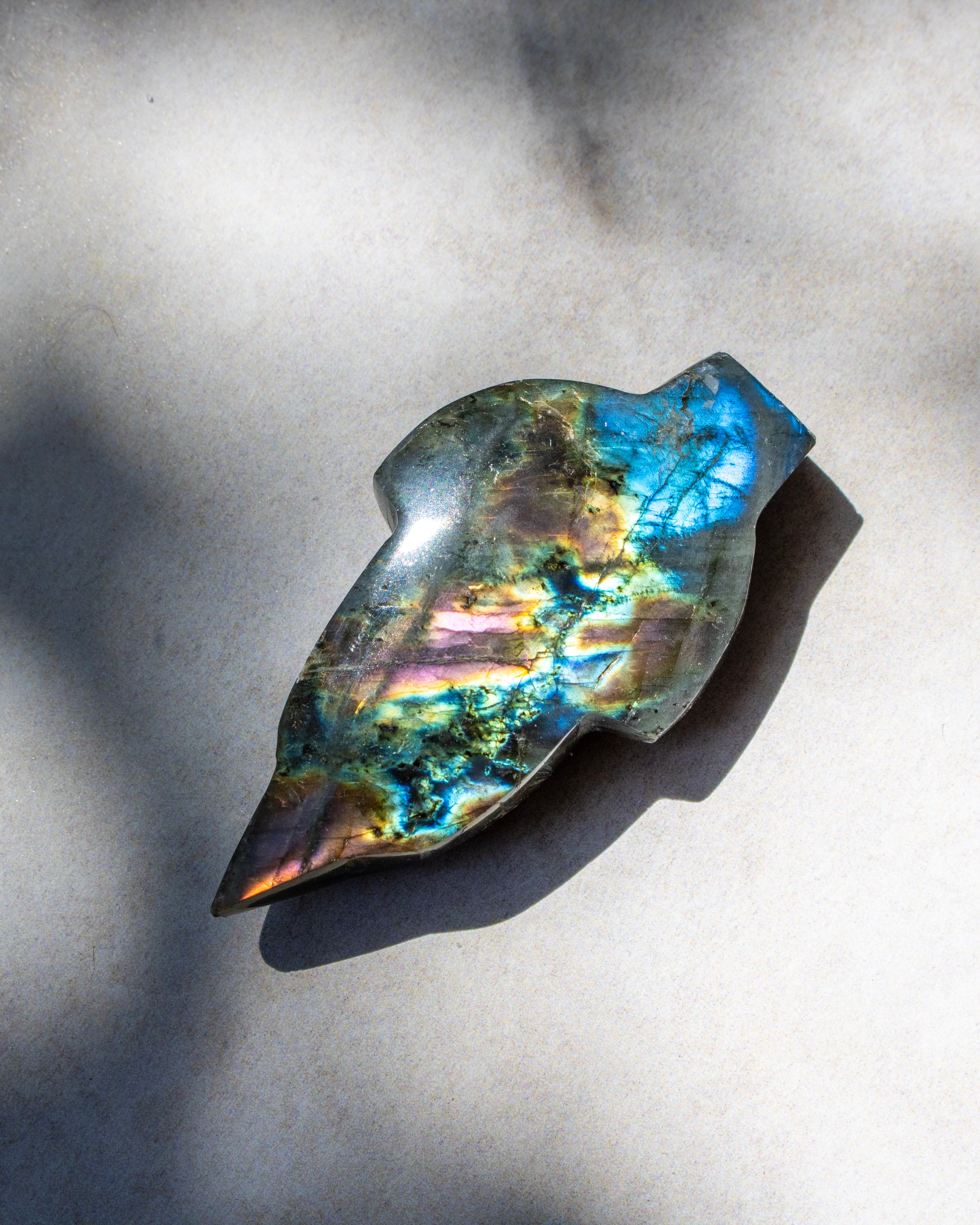 A polished labradorite stone cabochon with vibrant blue, green, and gold iridescence, resting on a textured gray surface under direct sunlight.