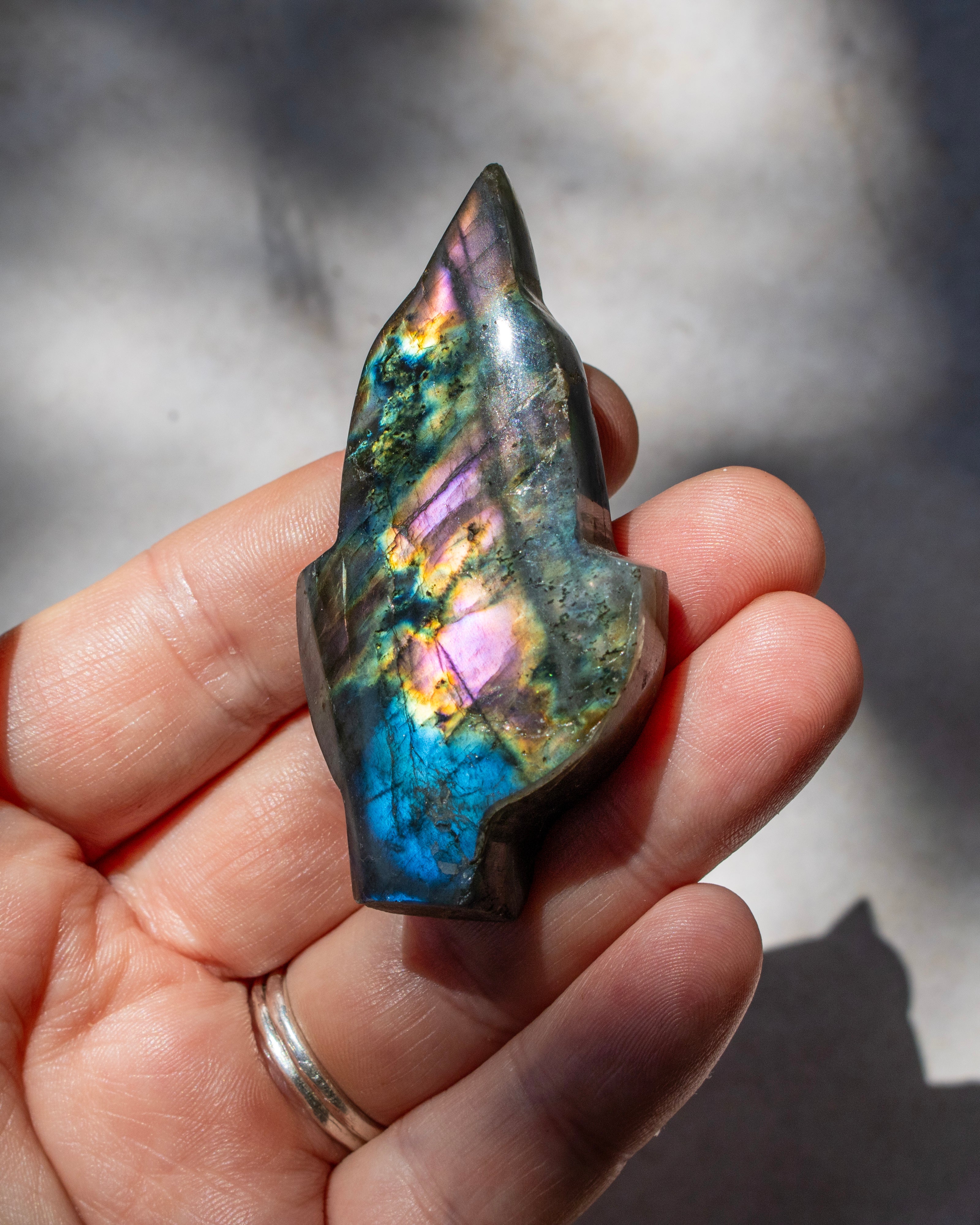 A hand holding a polished labradorite crystal point, showcasing its iridescent rainbow sheen with blue, purple, green, and gold colors, set against a soft-focus gray background with natural shadows.