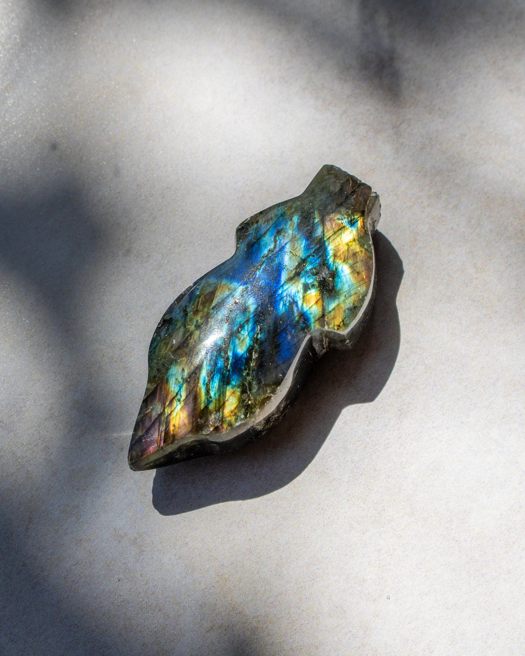 A raw labradorite gemstone with vibrant blue, green, and gold iridescence, resting on a light gray surface under direct sunlight with soft shadows.