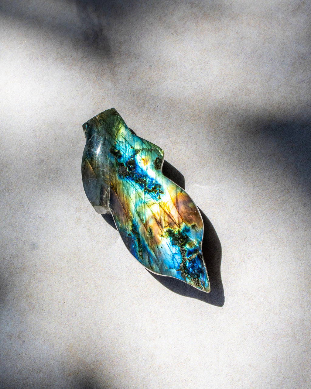 A polished labradorite stone cabochon with vibrant blue, green, gold, and brown iridescent flashes, displayed on a textured light gray surface under natural dappled sunlight.