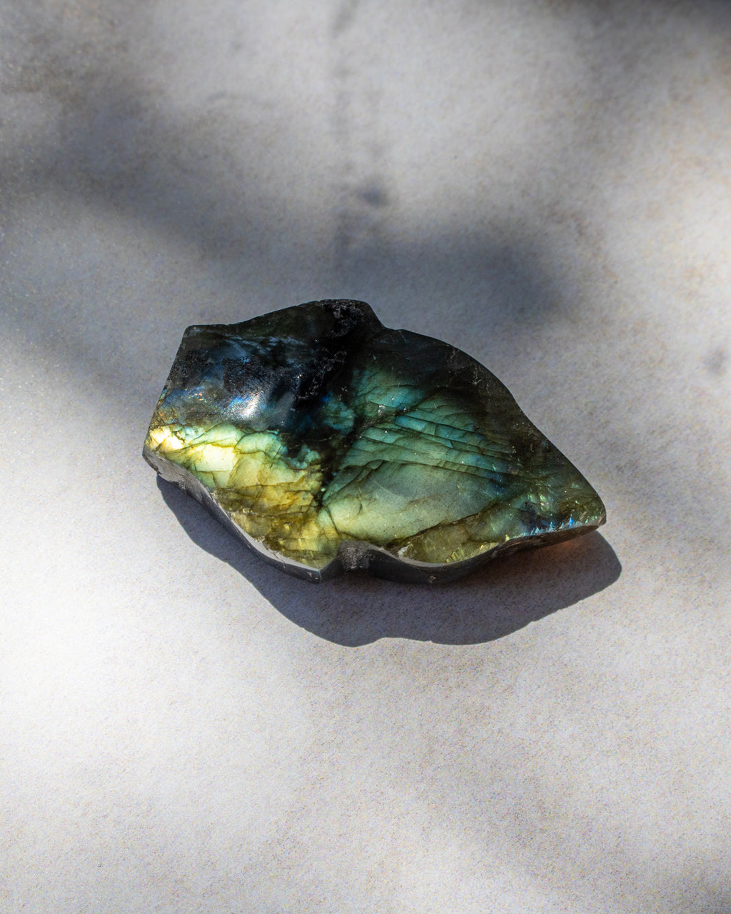 A raw labradorite crystal stone with a natural rough shape, displaying a shimmering green and gold iridescent finish, resting on a light gray textured surface with soft shadows.