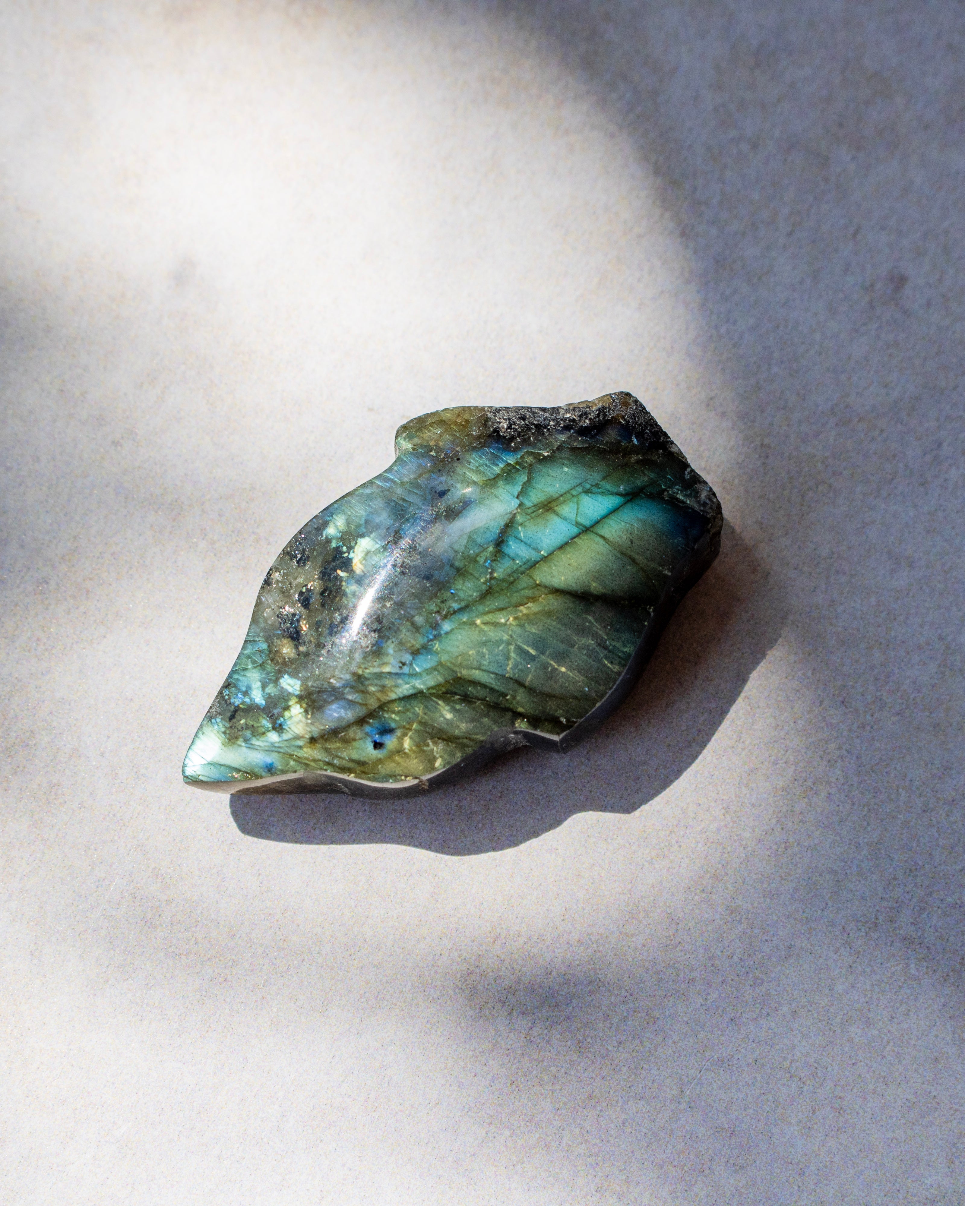 A raw labradorite crystal stone with a shimmering green and blue iridescent finish, resting on a neutral gray surface with soft shadows.