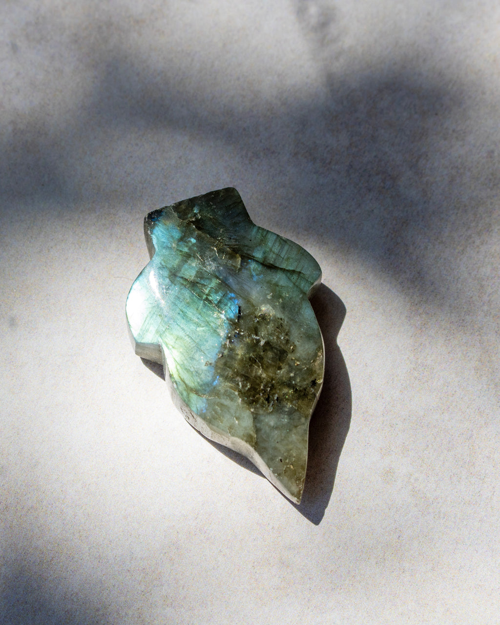 A polished natural crystal or gemstone with a pear-shaped form, displaying iridescent green, blue, and gold labradorescence, resting on a light gray surface under direct sunlight.