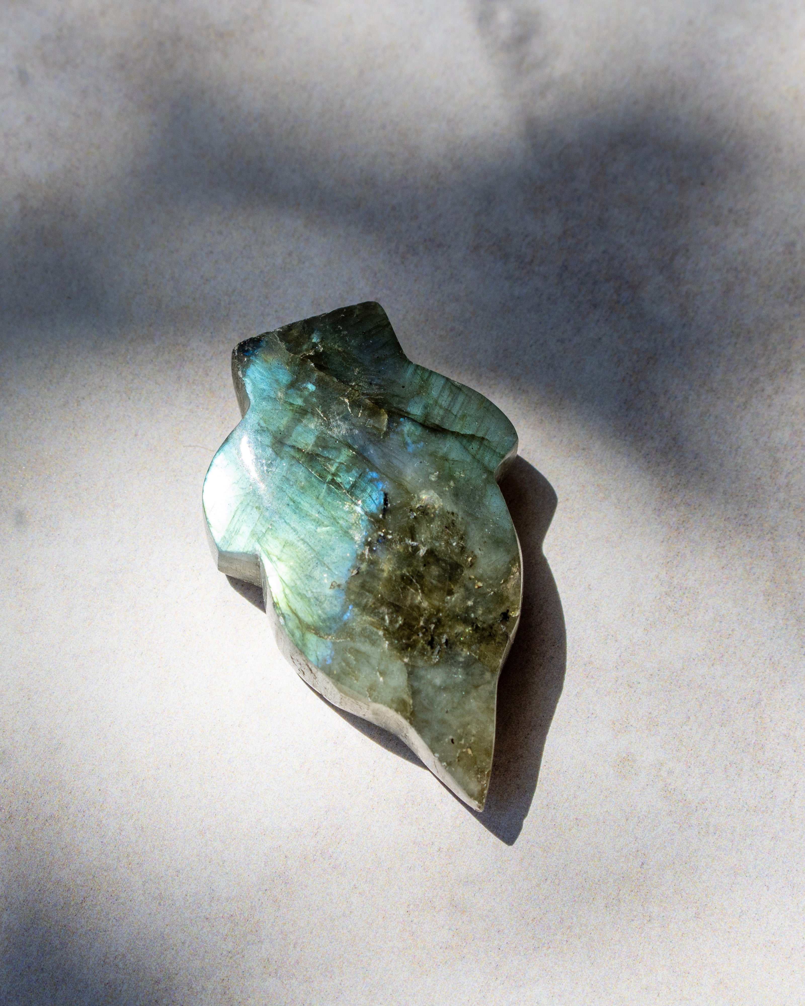 A polished natural crystal or gemstone with a pear-shaped form, displaying iridescent green, blue, and gold labradorescence, resting on a light gray surface under direct sunlight.