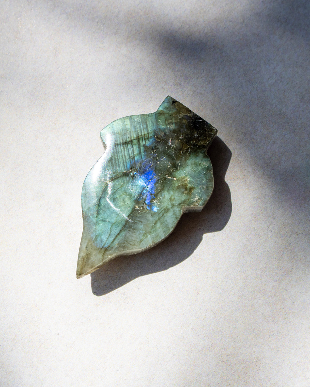 A polished labradorite stone pendant with a leaf-like shape, displaying natural green and blue flashes, resting on a neutral light gray background with soft shadows.