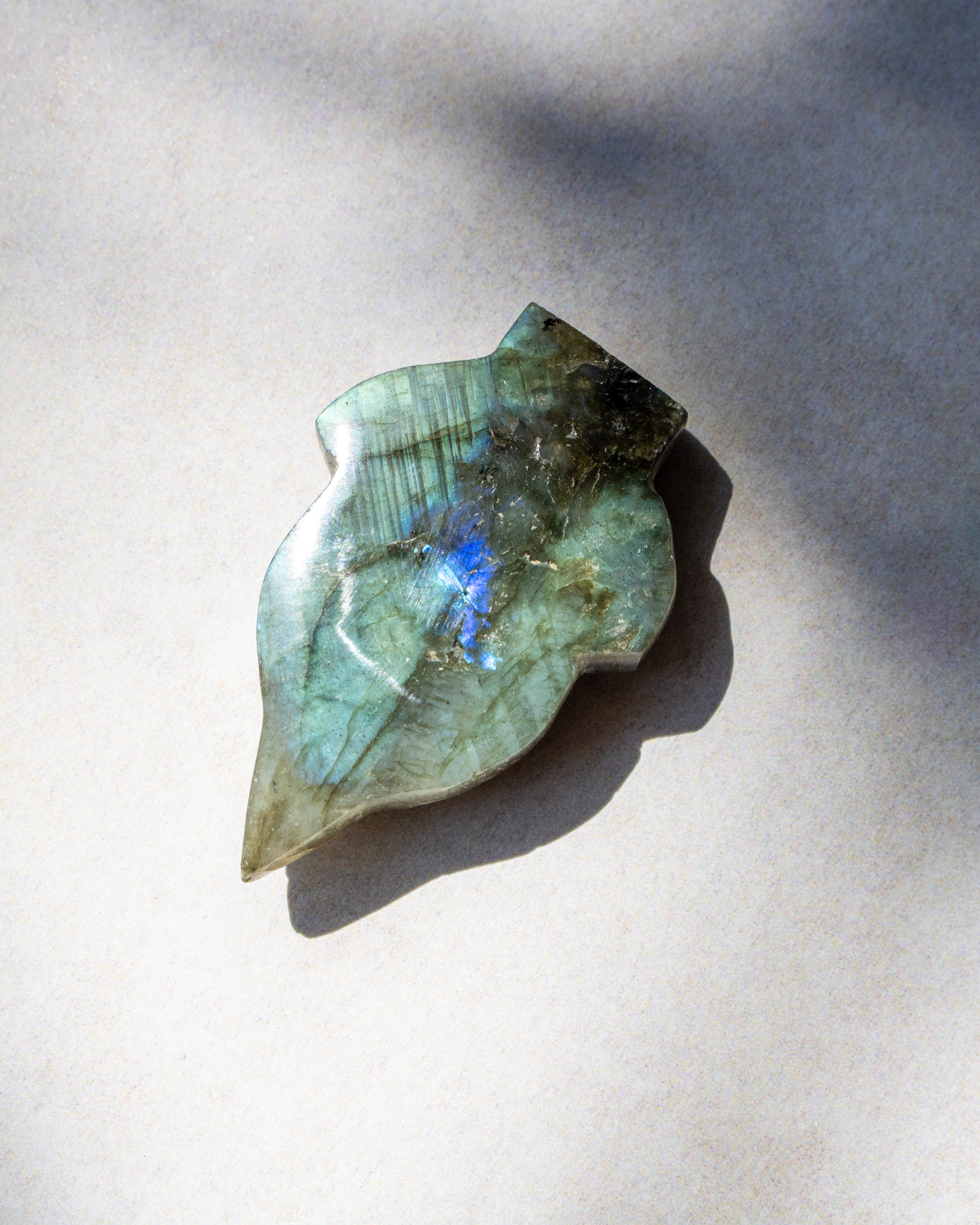 A polished labradorite stone pendant with a leaf-like shape, displaying natural green and blue flashes, resting on a neutral light gray background with soft shadows.
