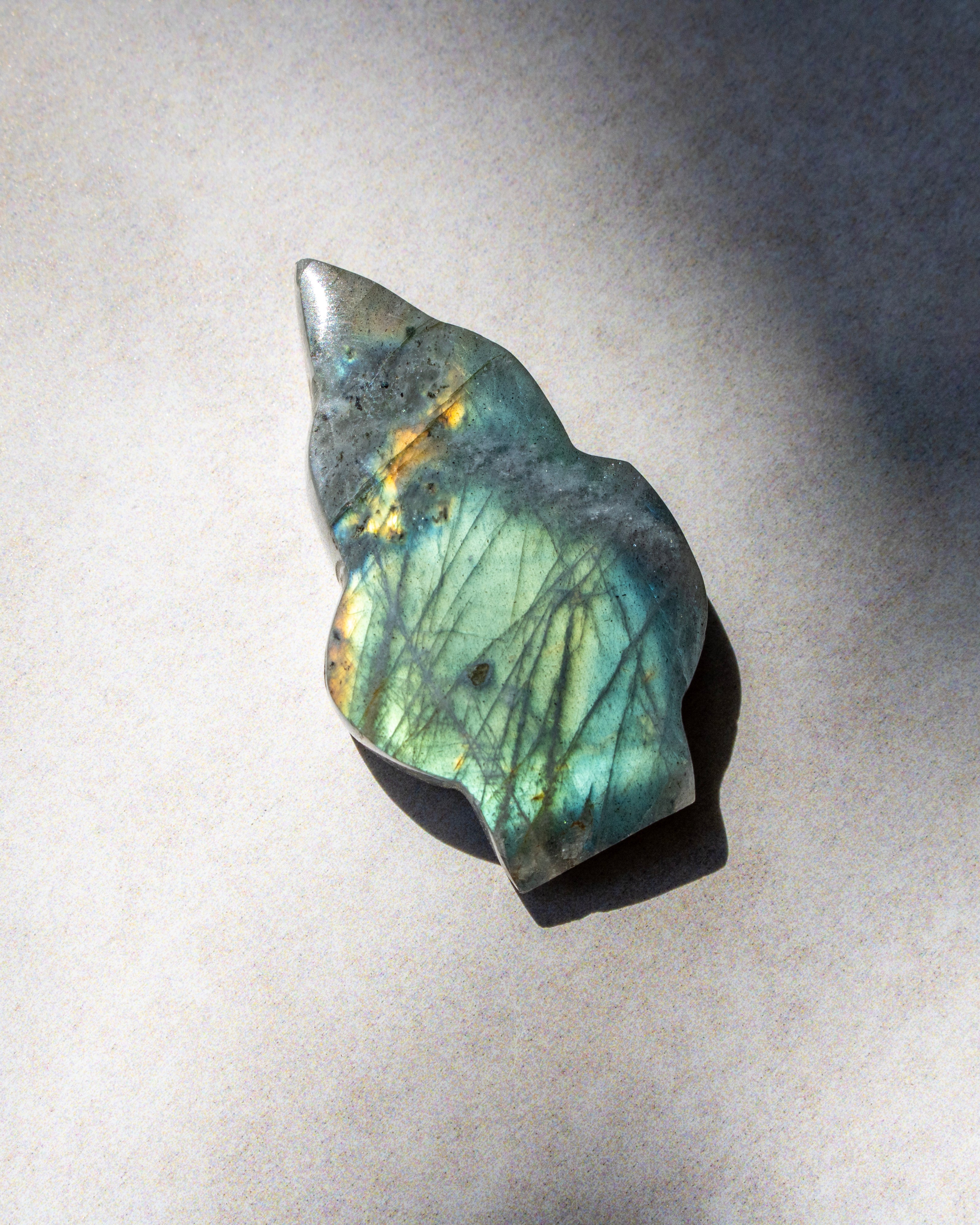 A polished labradorite stone pendant with a natural irregular shape, displaying a shimmering green and blue iridescent surface with fine gold and brown crystalline structures, resting on a neutral light gray background.