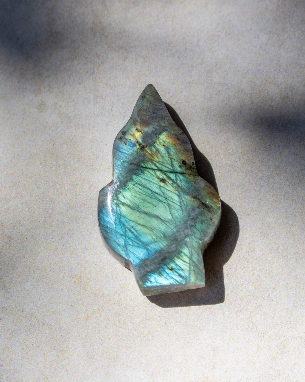 A polished labradorite crystal stone with a natural irregular shape, displaying a shimmering blue-green color with gold and brown inclusions, resting on a neutral light gray surface.