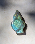 A polished labradorite crystal stone with a natural irregular shape, displaying a shimmering blue-green color with gold and brown inclusions, resting on a neutral light gray surface.