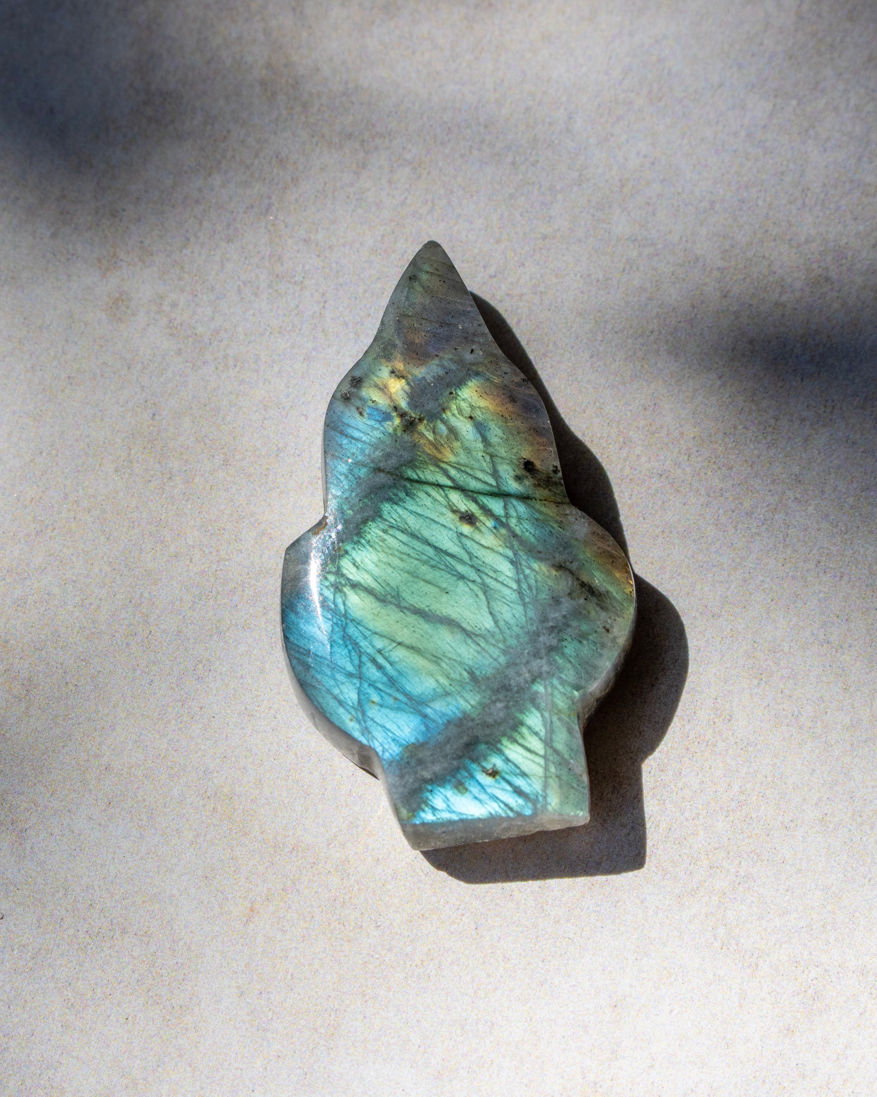 A polished labradorite crystal stone with a natural irregular shape, displaying a shimmering blue-green color with gold and brown inclusions, resting on a neutral light gray surface.