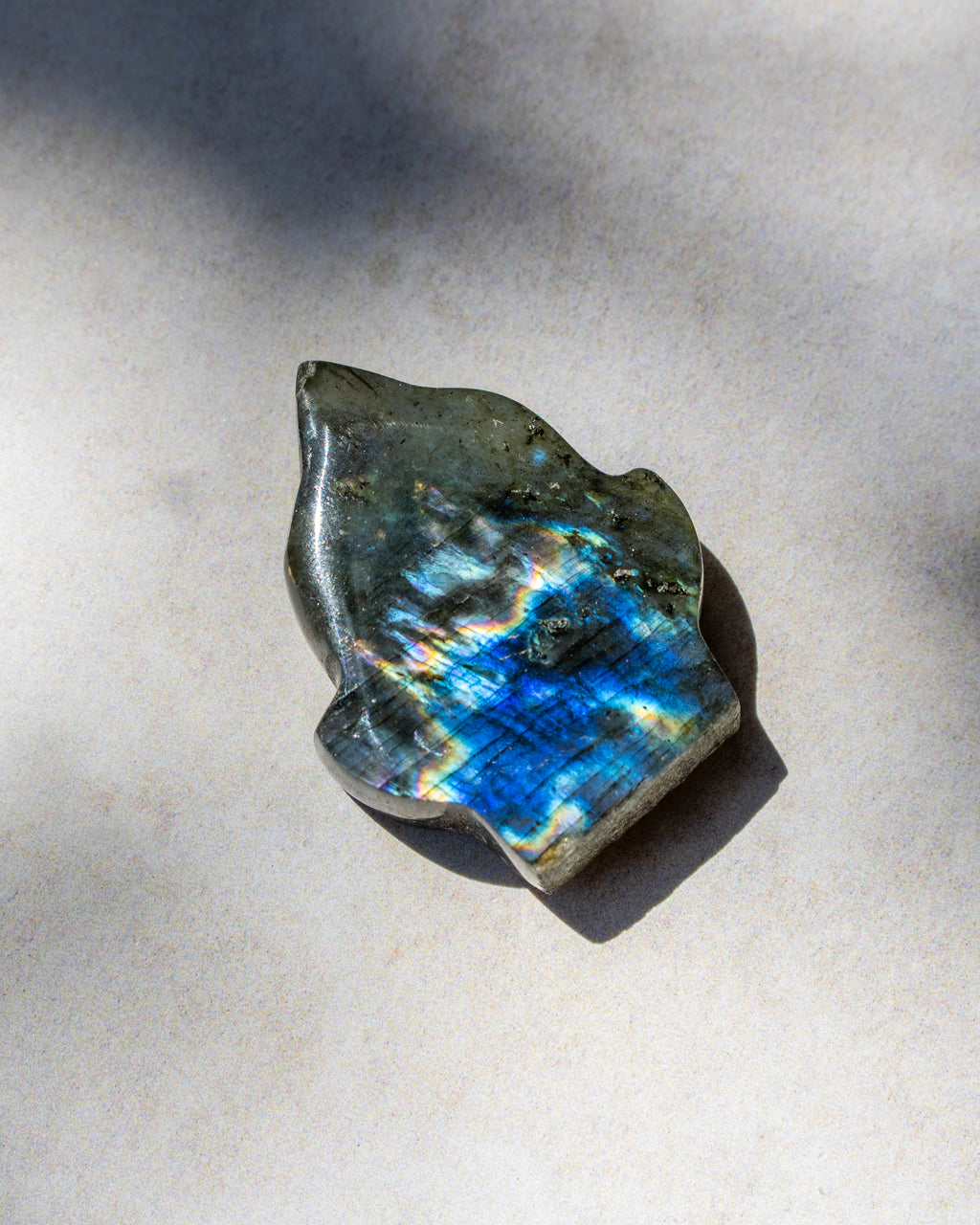 A polished irregular stone with a dark gray base and vivid blue iridescent flashes, photographed on a light neutral surface with strong sunlight and shadows.