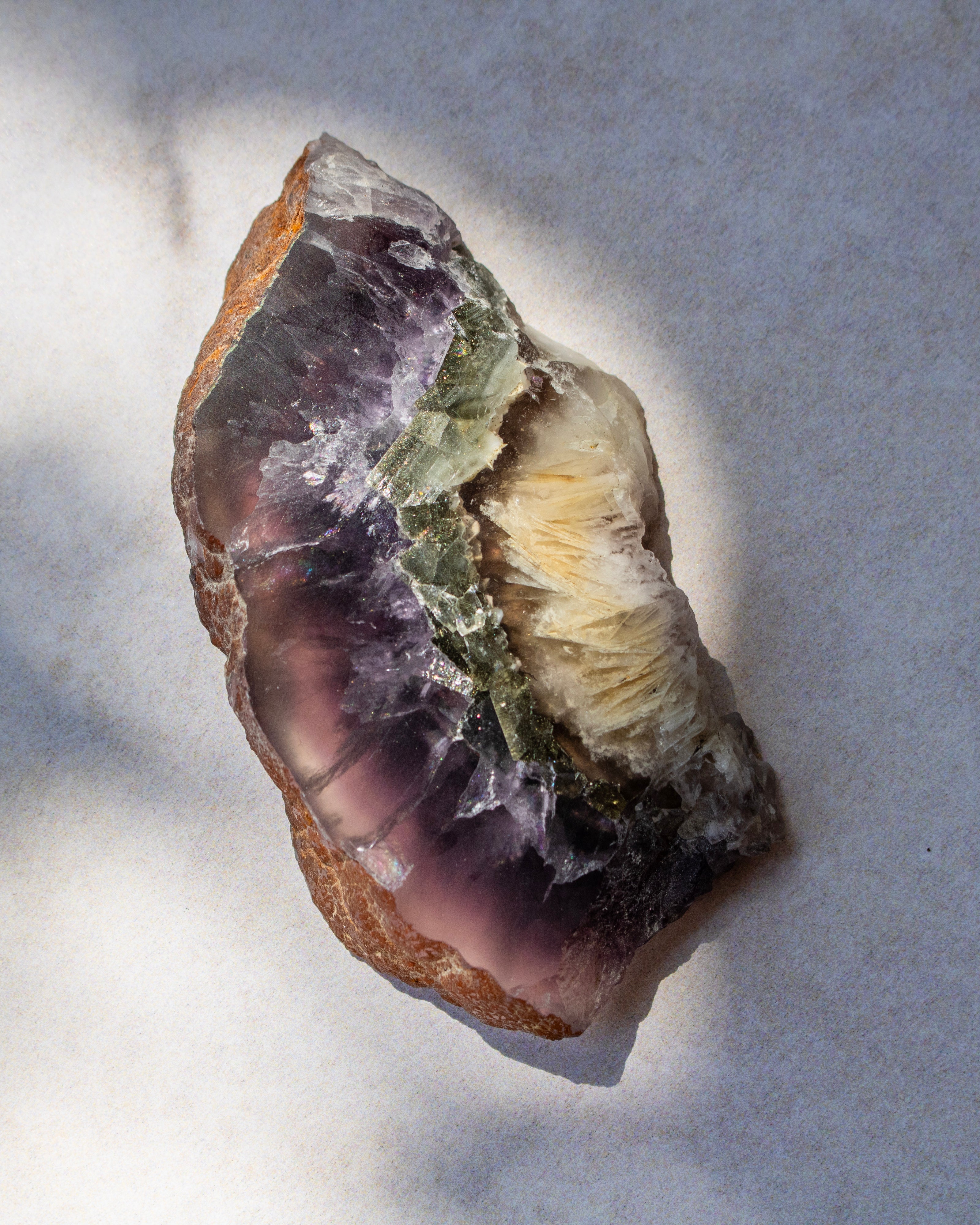 A raw purple geode slice with a natural rough outer crust, showing a purple crystal cavity filled with white and light green quartz, photographed on a light gray textured surface.