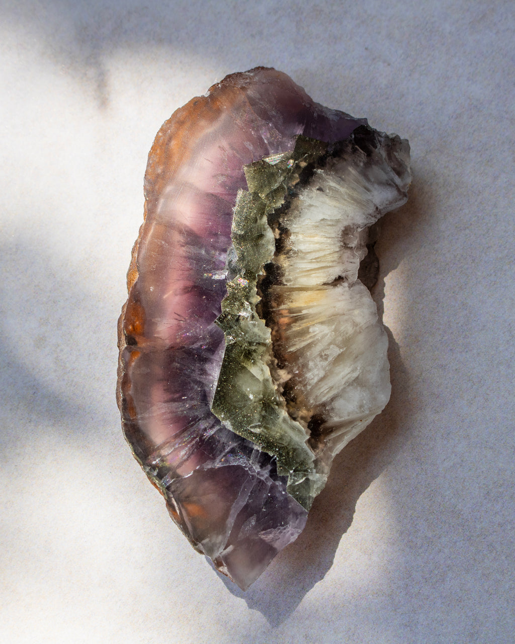 A raw mineral specimen featuring layered bands of purple fluorite, translucent brown, and a central greenish crystalline section, resting on a light gray surface.