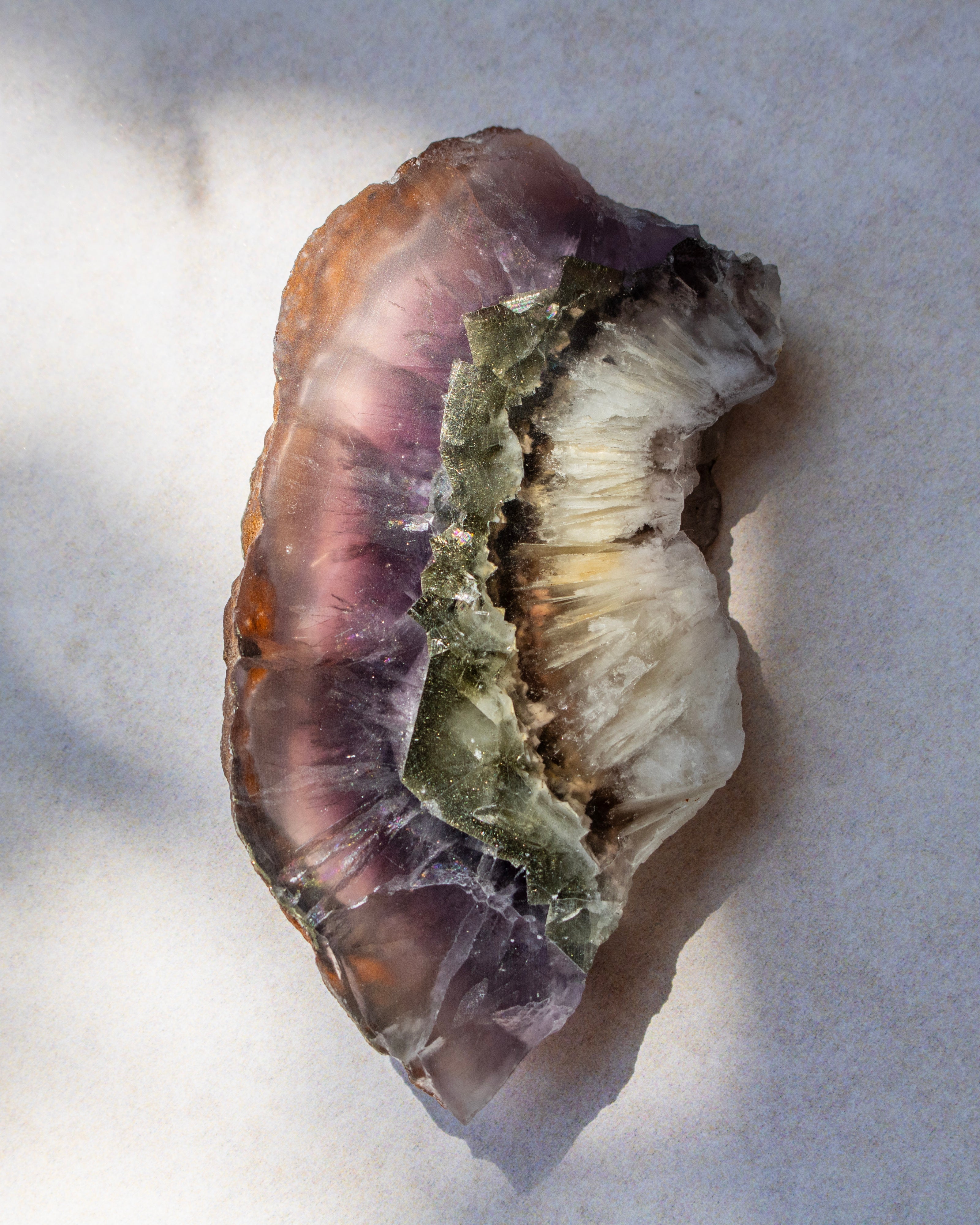 A raw mineral specimen featuring layered bands of purple fluorite, translucent brown, and a central greenish crystalline section, resting on a light gray surface.