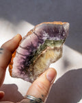A hand holding a raw purple geode slice with visible layers of purple, green, and cream quartz, set against a neutral gray background with soft shadows.
