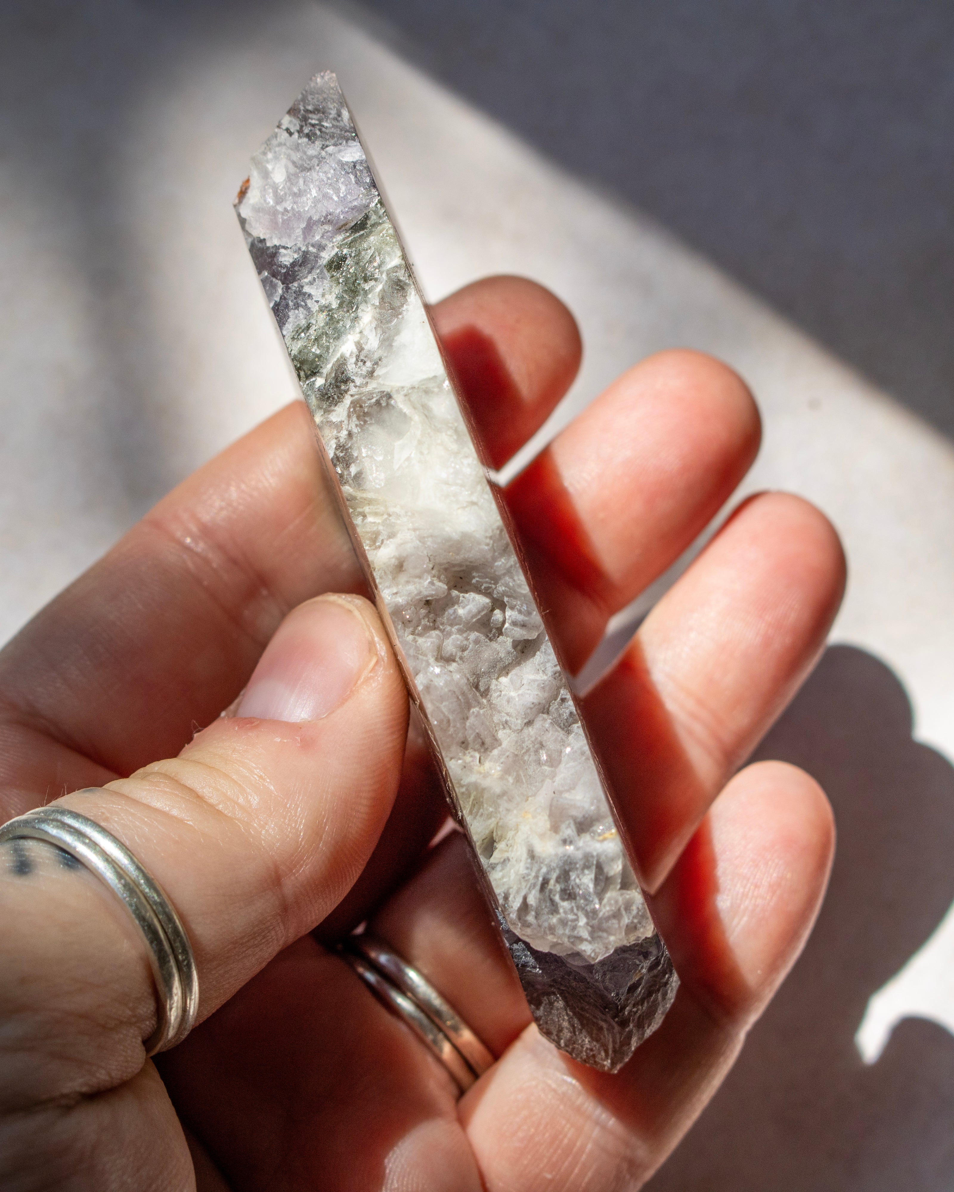 A hand holding a raw quartz crystal point wand under direct sunlight, showing its natural gray and white crystalline texture.