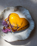 A polished yellow heart-shaped stone or glass ornament with natural marbled veining, displayed on a white speckled ceramic dish with dried purple flowers on a neutral surface.
