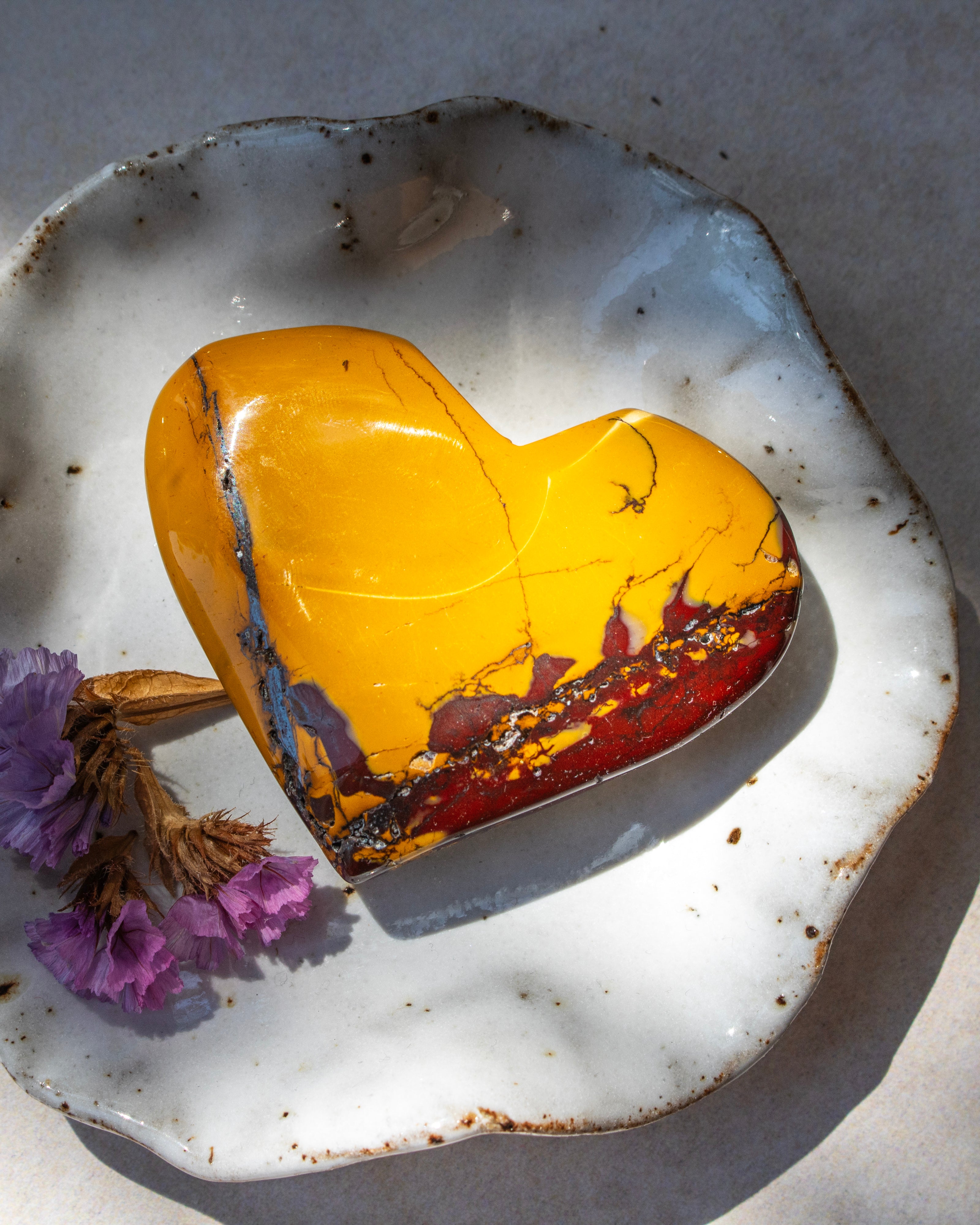 A heart-shaped stone with a yellow top and a red-brown bottom, displayed on a rustic white ceramic dish with dried purple flowers.