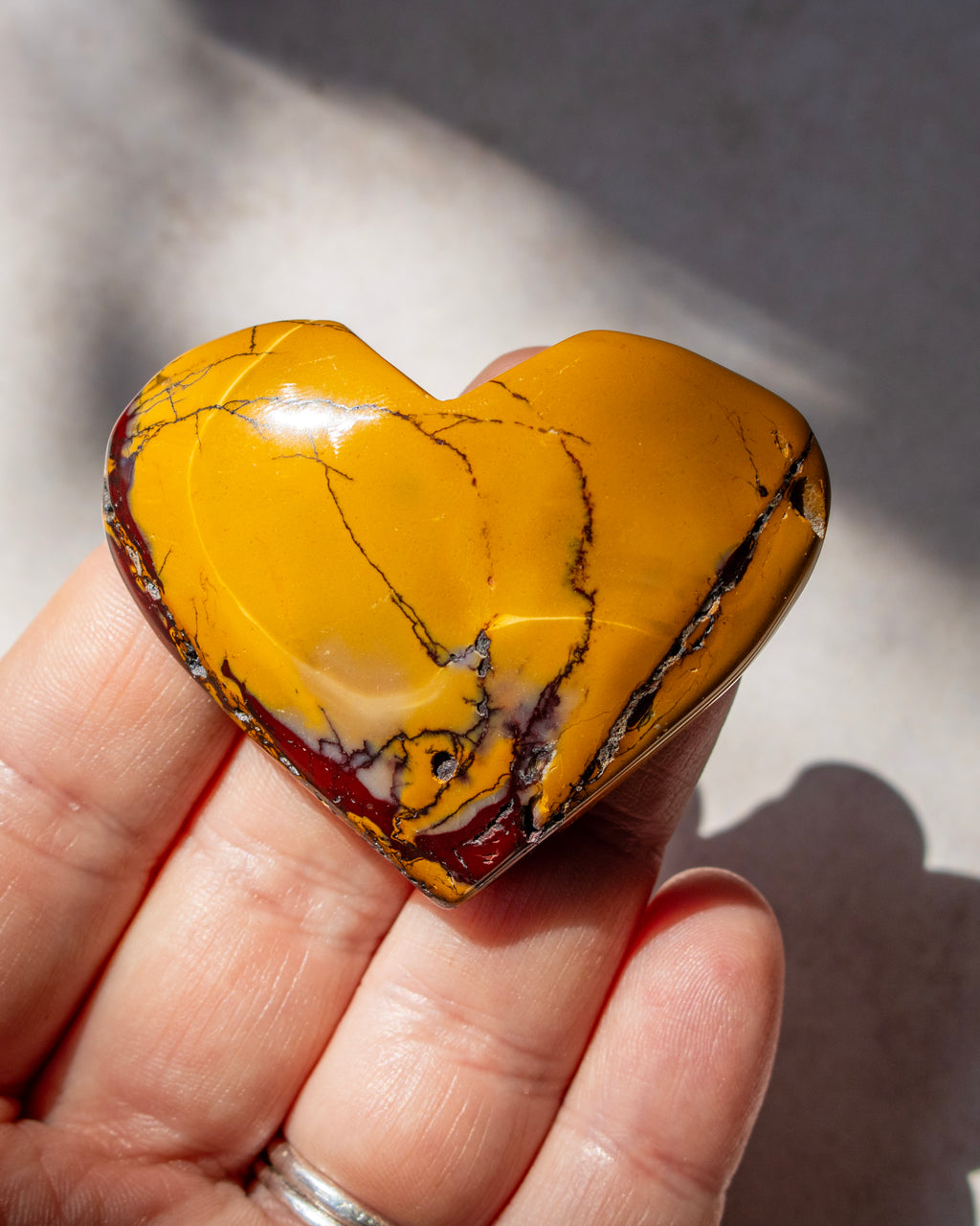 A heart-shaped polished stone with a golden yellow body and dark brown to black veining, held in a person's hand against a neutral light gray background.