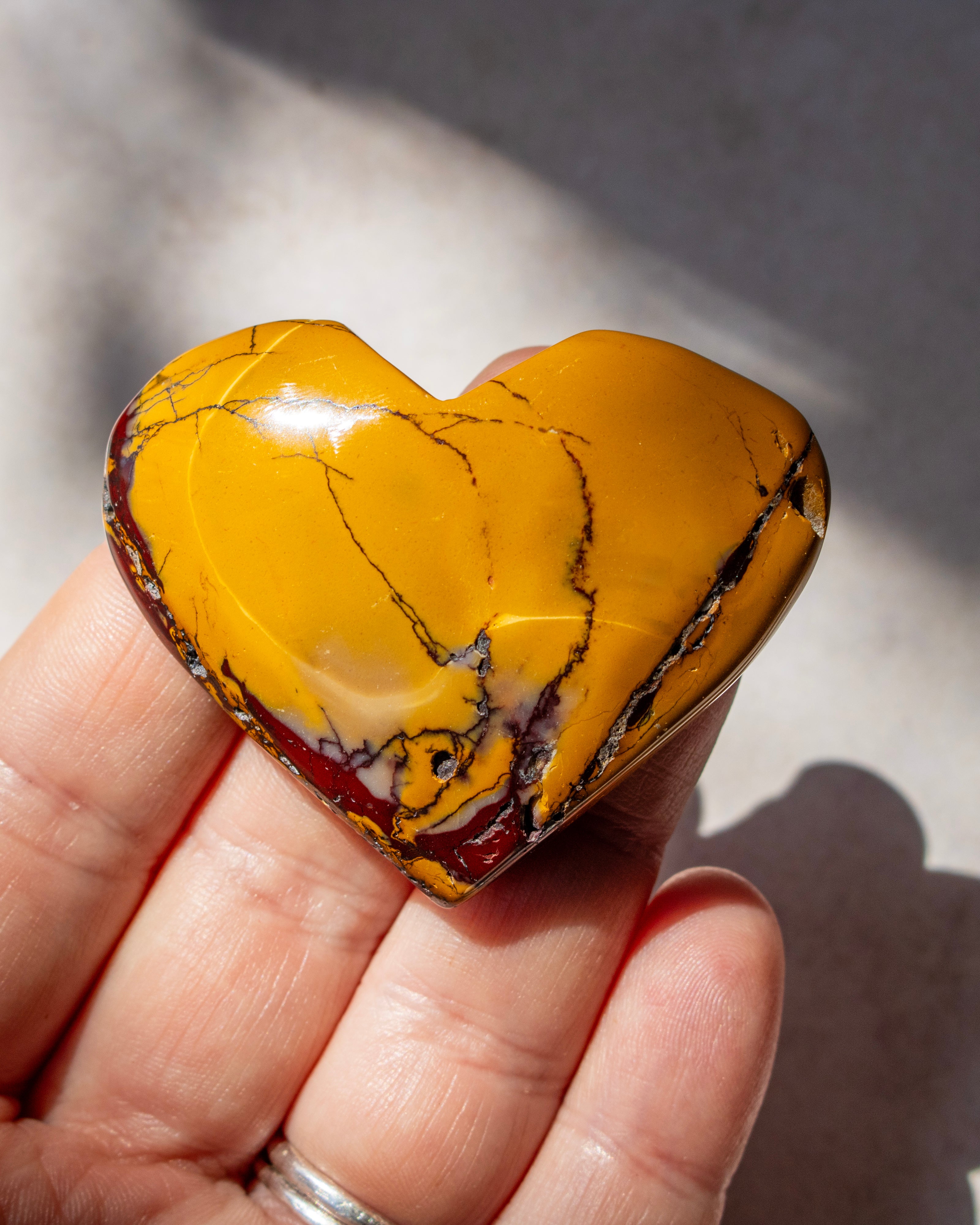 A heart-shaped polished stone with a golden yellow body and dark brown to black veining, held in a person's hand against a neutral light gray background.