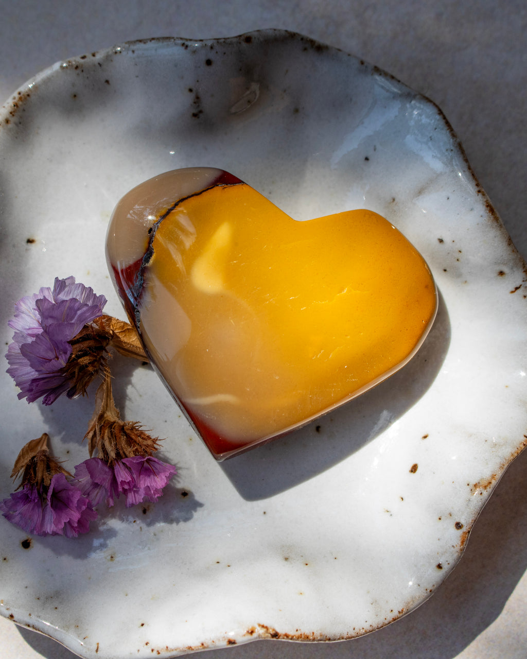 A heart-shaped stone with a vibrant yellow and amber center and a clear outer shell, resting on a white speckled ceramic dish with dried purple flowers.