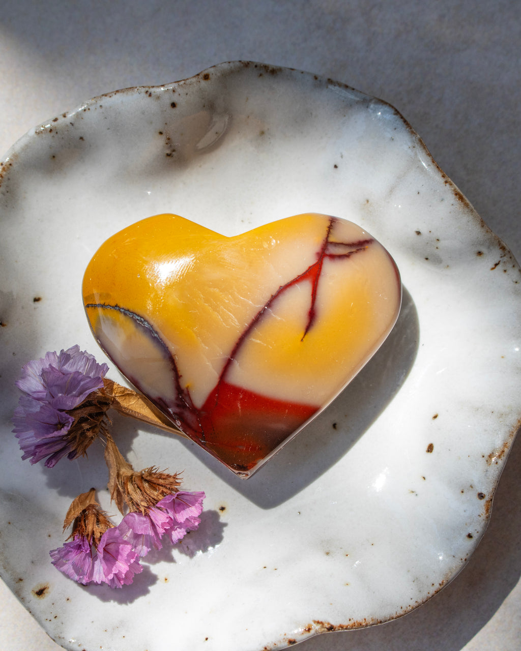 A heart-shaped  stone ornament with yellow, white, and red marbled patterns, displayed on a rustic white ceramic dish with dried purple flowers.