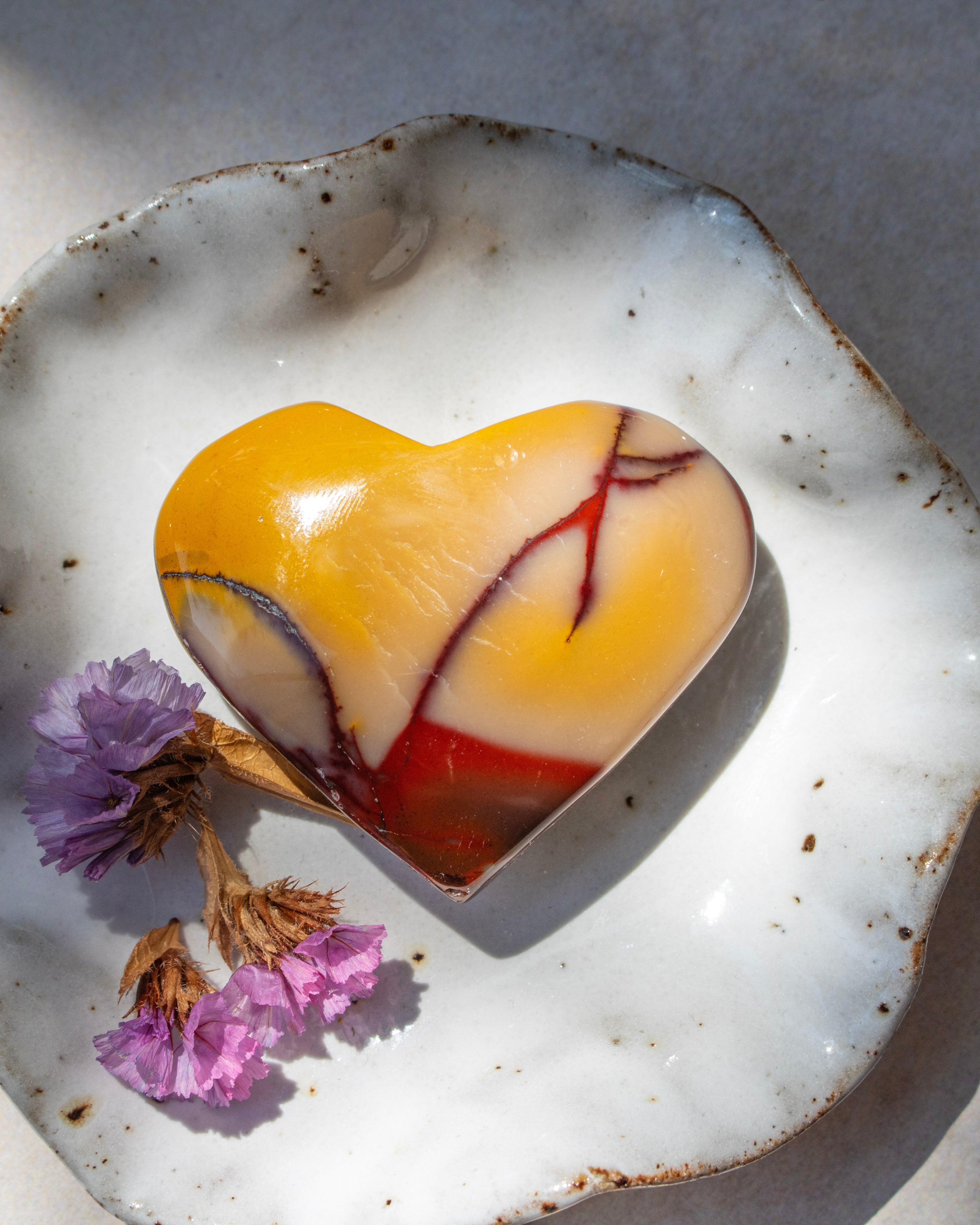 A heart-shaped  stone ornament with yellow, white, and red marbled patterns, displayed on a rustic white ceramic dish with dried purple flowers.