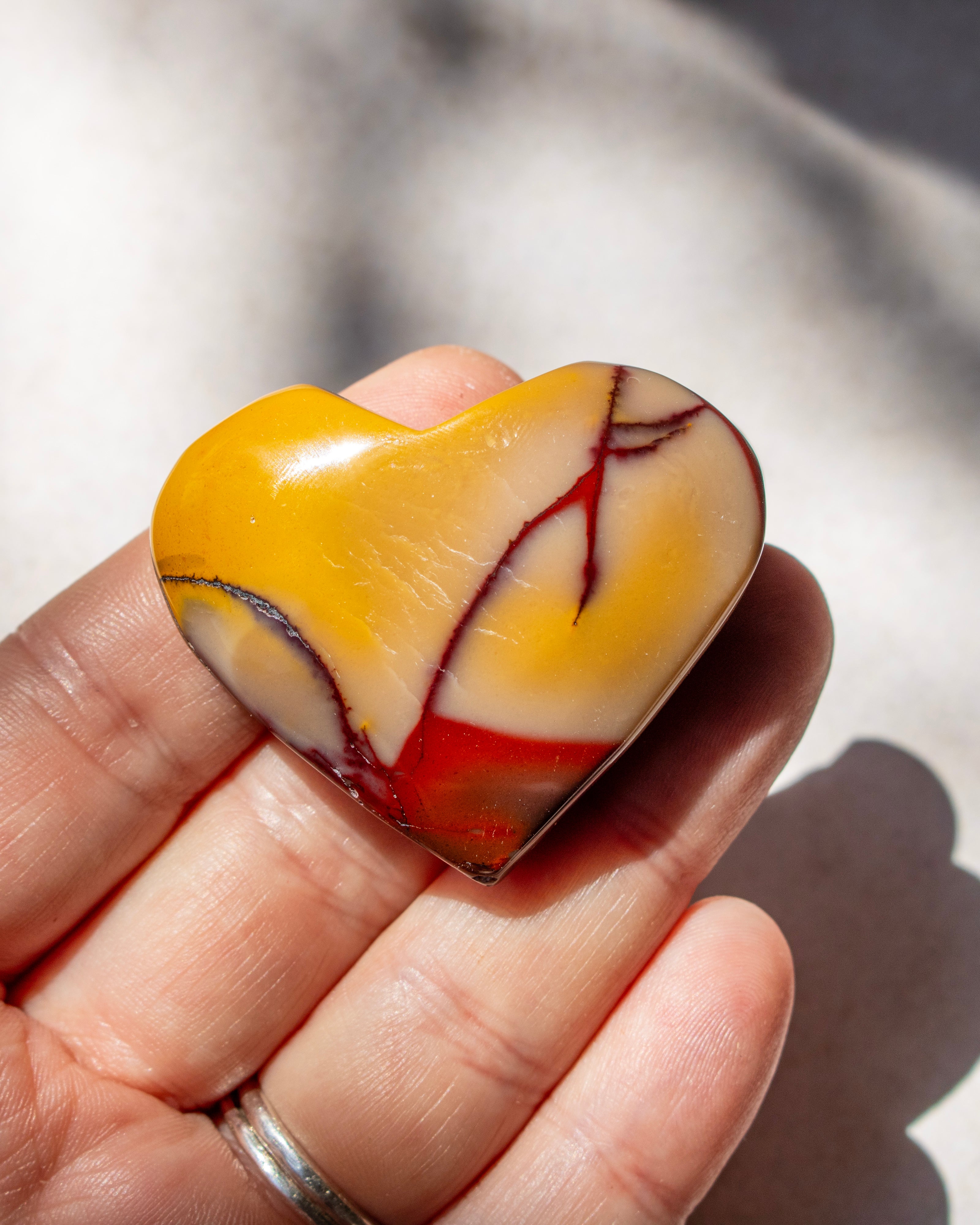 A heart-shaped polished stone with yellow, cream, and red marbled patterns, held in a person's hand under natural sunlight.