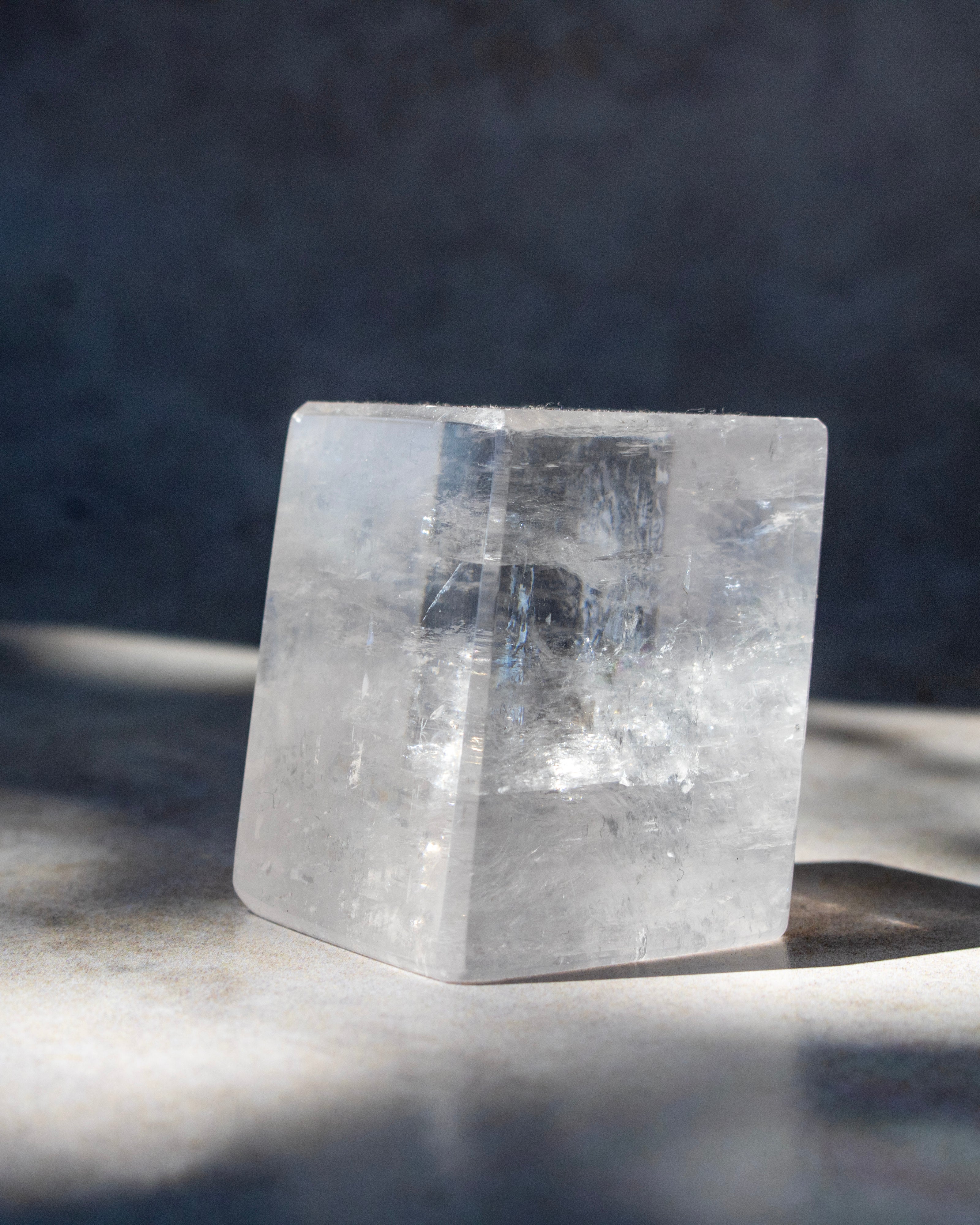 A clear crystal cube with natural inclusions, photographed on a textured gray surface with dramatic side lighting and soft shadows.