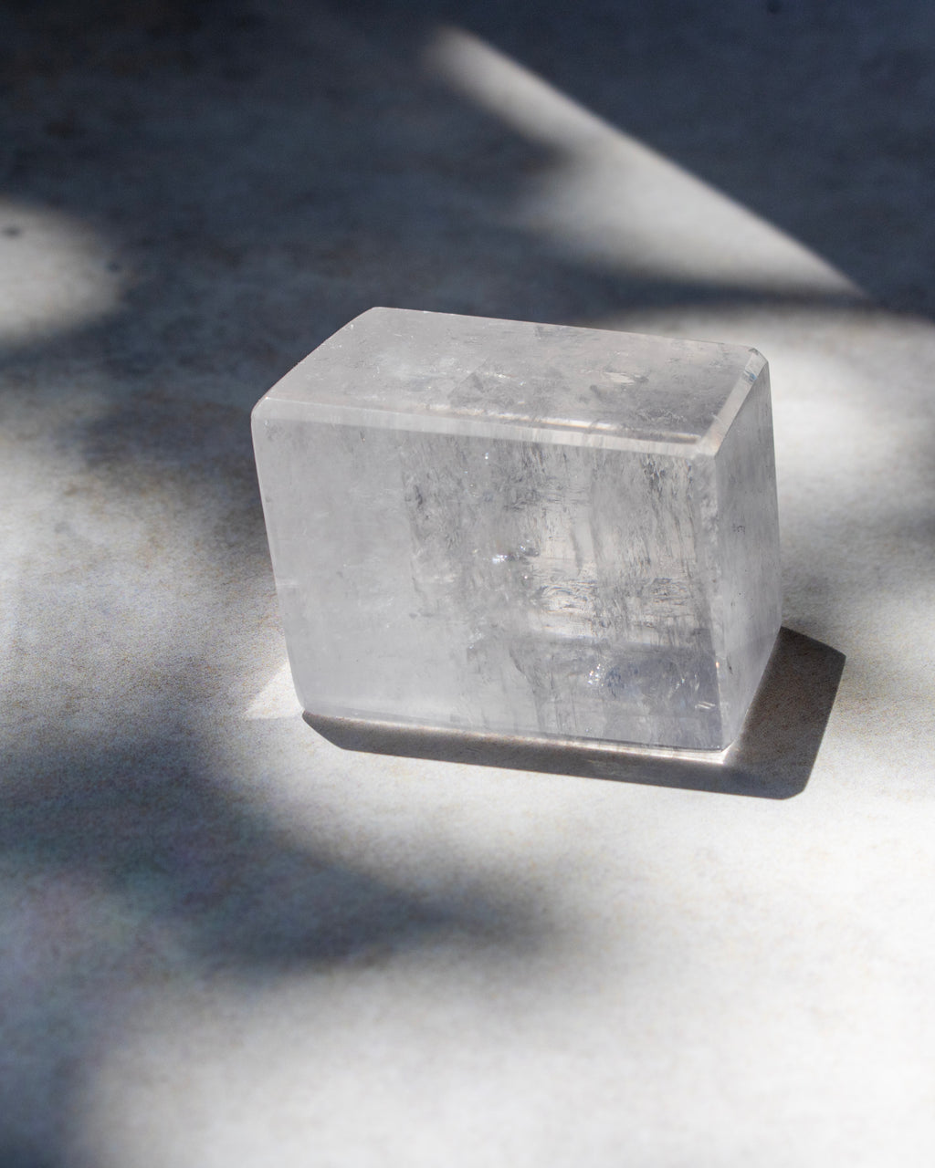 A clear rectangular crystal block, likely quartz, resting on a gray surface with dramatic natural shadows.