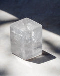 A clear, faceted crystal cube with natural internal textures, resting on a light gray surface under direct sunlight that casts a sharp shadow.