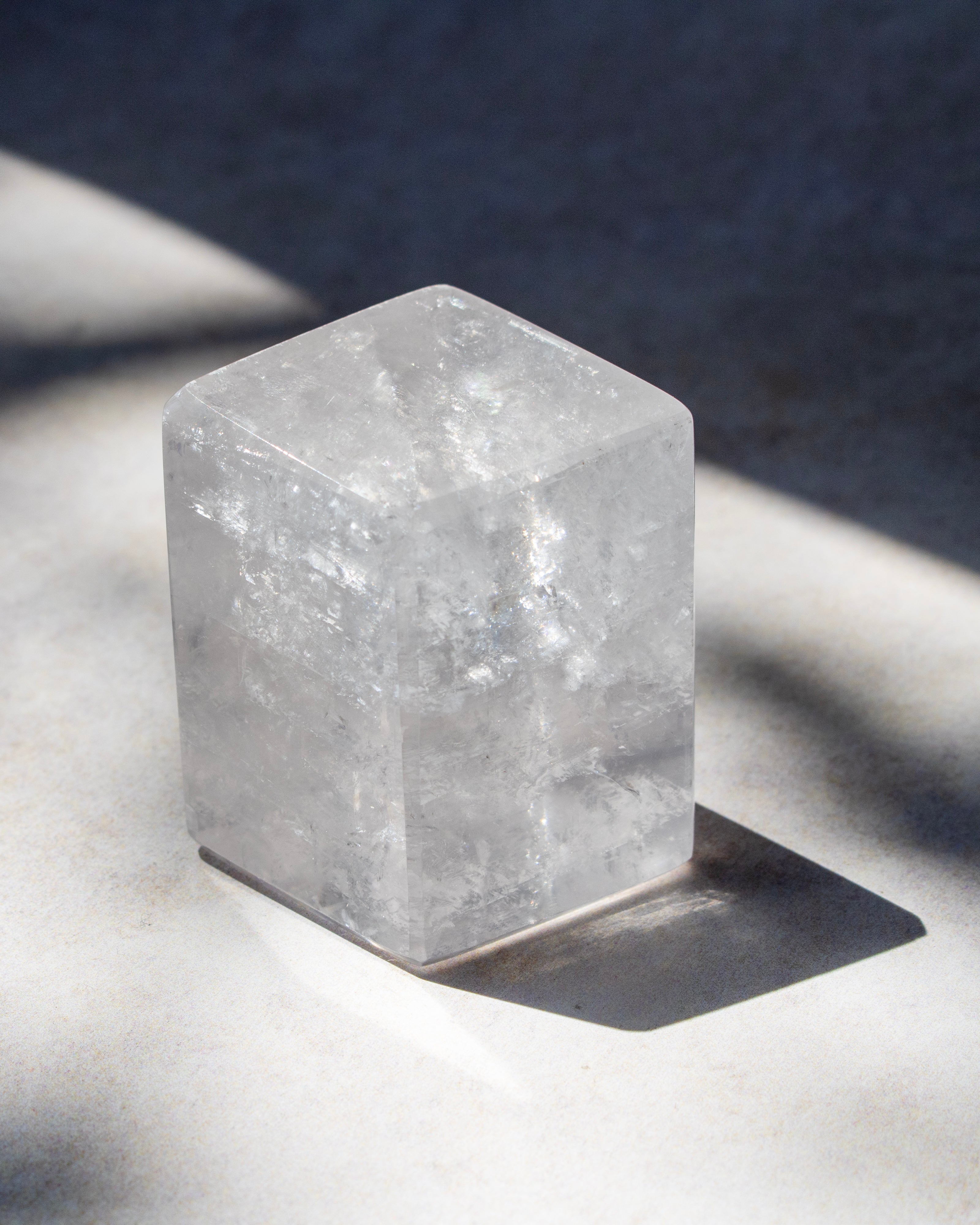 A clear, faceted crystal cube with natural internal textures, resting on a light gray surface under direct sunlight that casts a sharp shadow.