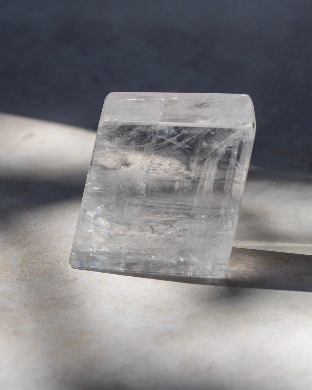 A clear rectangular crystal stone with visible internal linear inclusions, photographed on a light surface with strong directional shadows against a dark gray background.