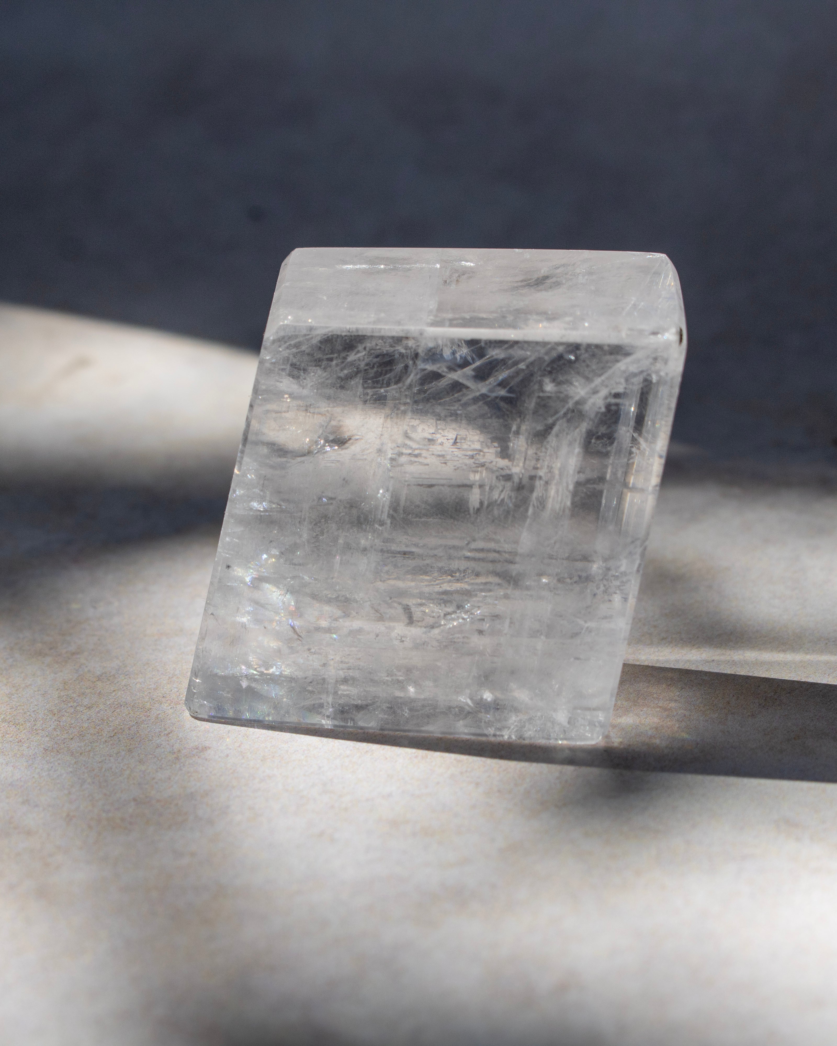 A clear rectangular crystal stone with visible internal linear inclusions, photographed on a light surface with strong directional shadows against a dark gray background.