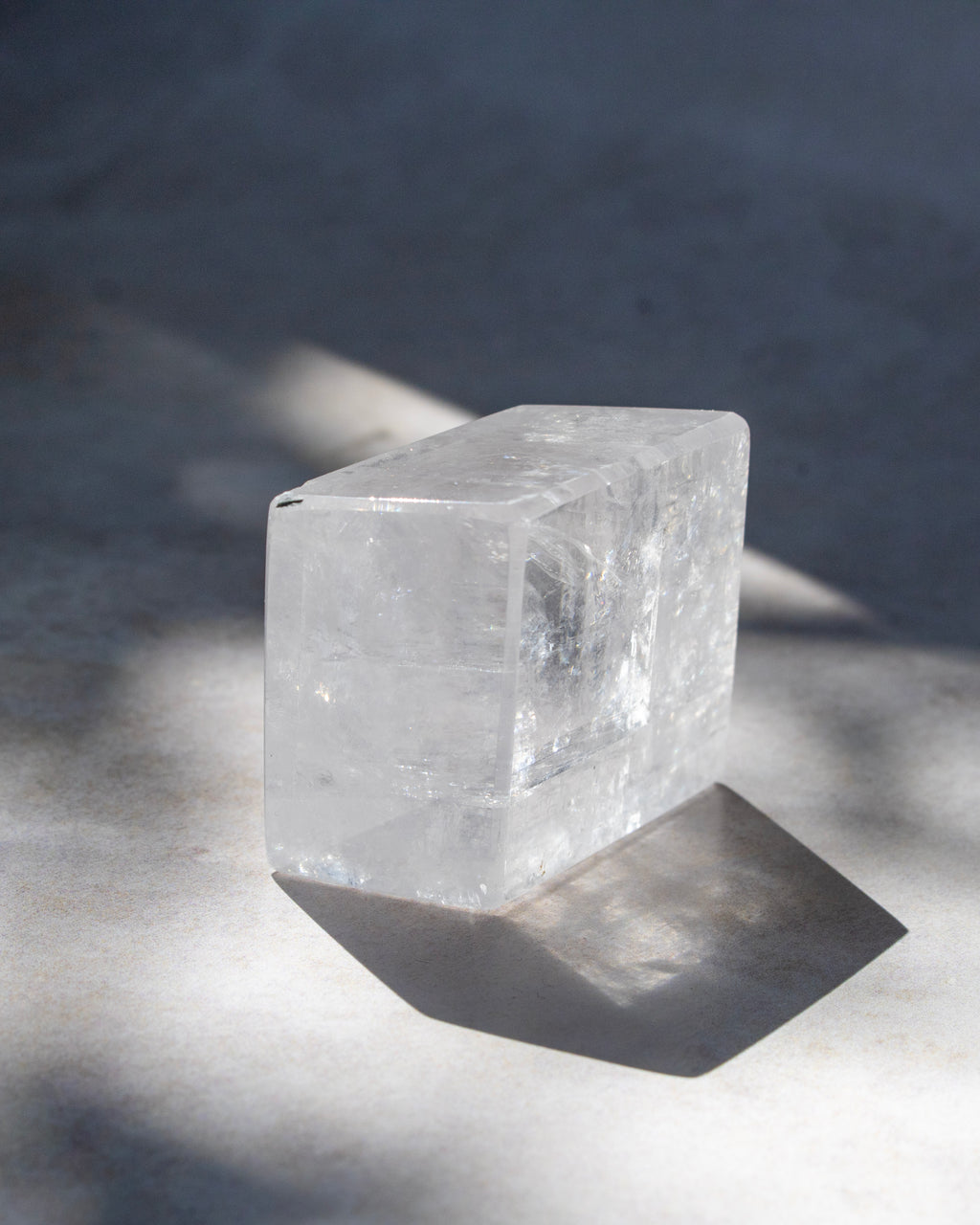 A clear rectangular crystal or stone block with a polished finish, photographed on a light surface with strong natural shadows.