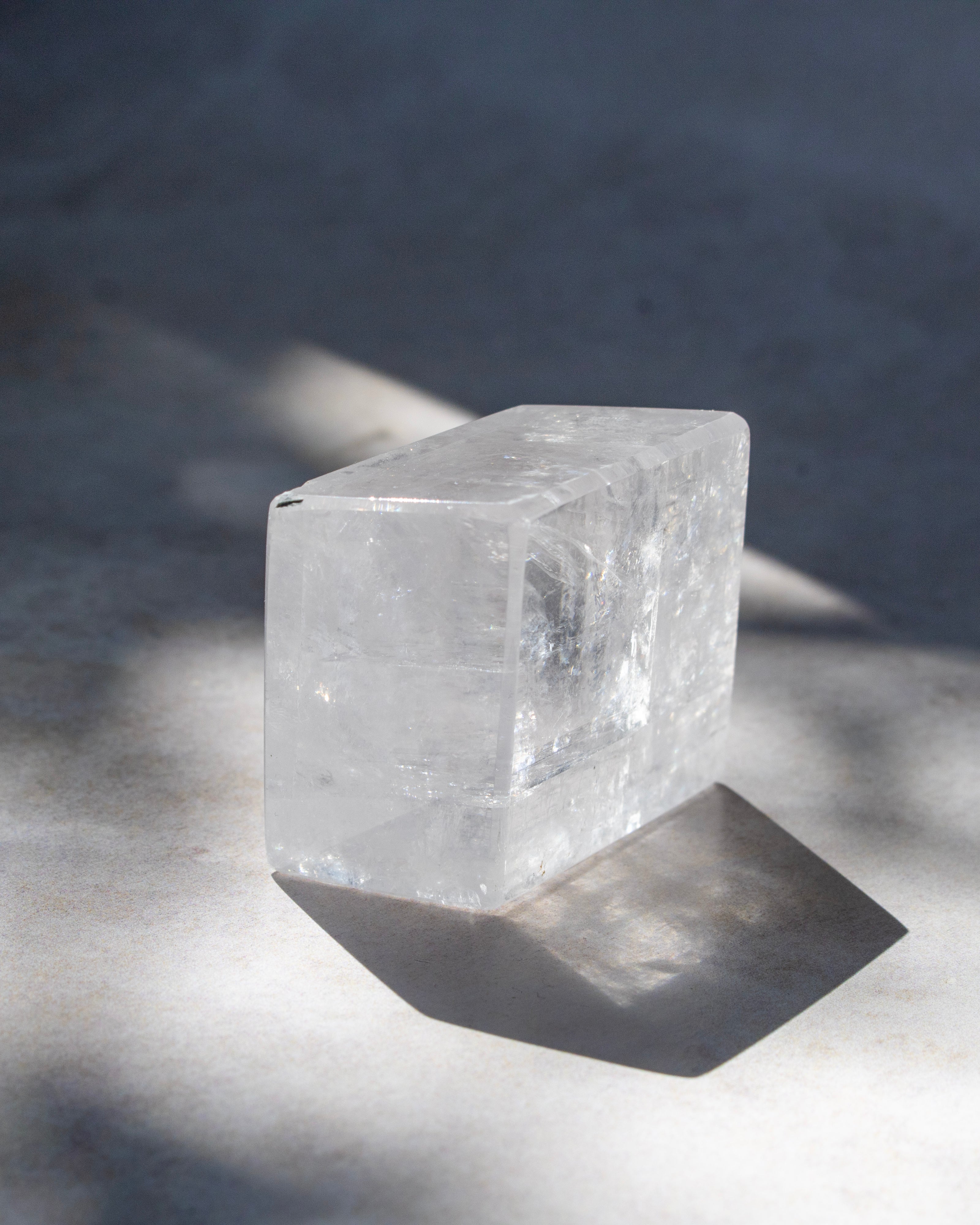A clear rectangular crystal or stone block with a polished finish, photographed on a light surface with strong natural shadows.