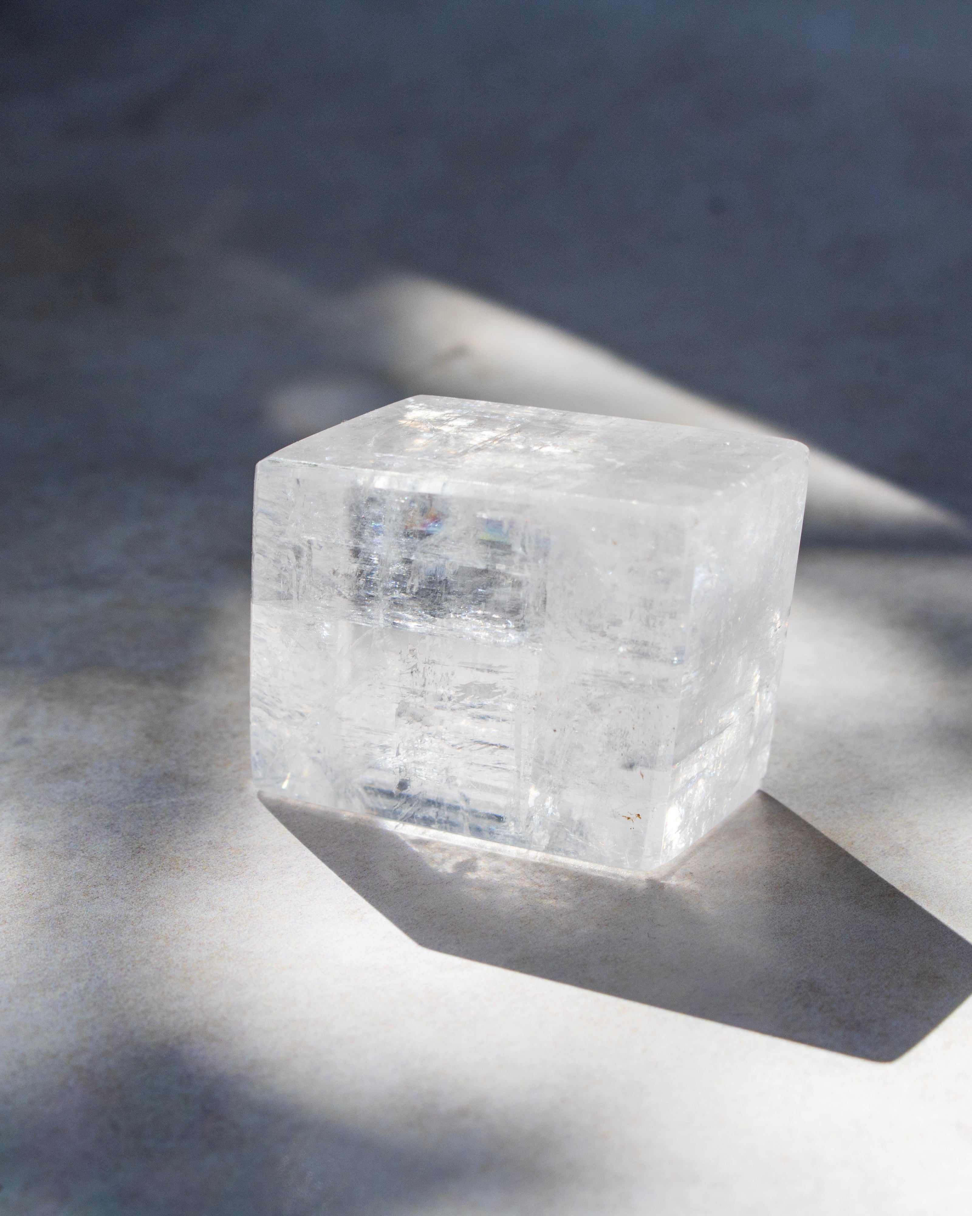 A clear, translucent cube-shaped crystal or stone block placed on a gray surface with strong natural shadows.