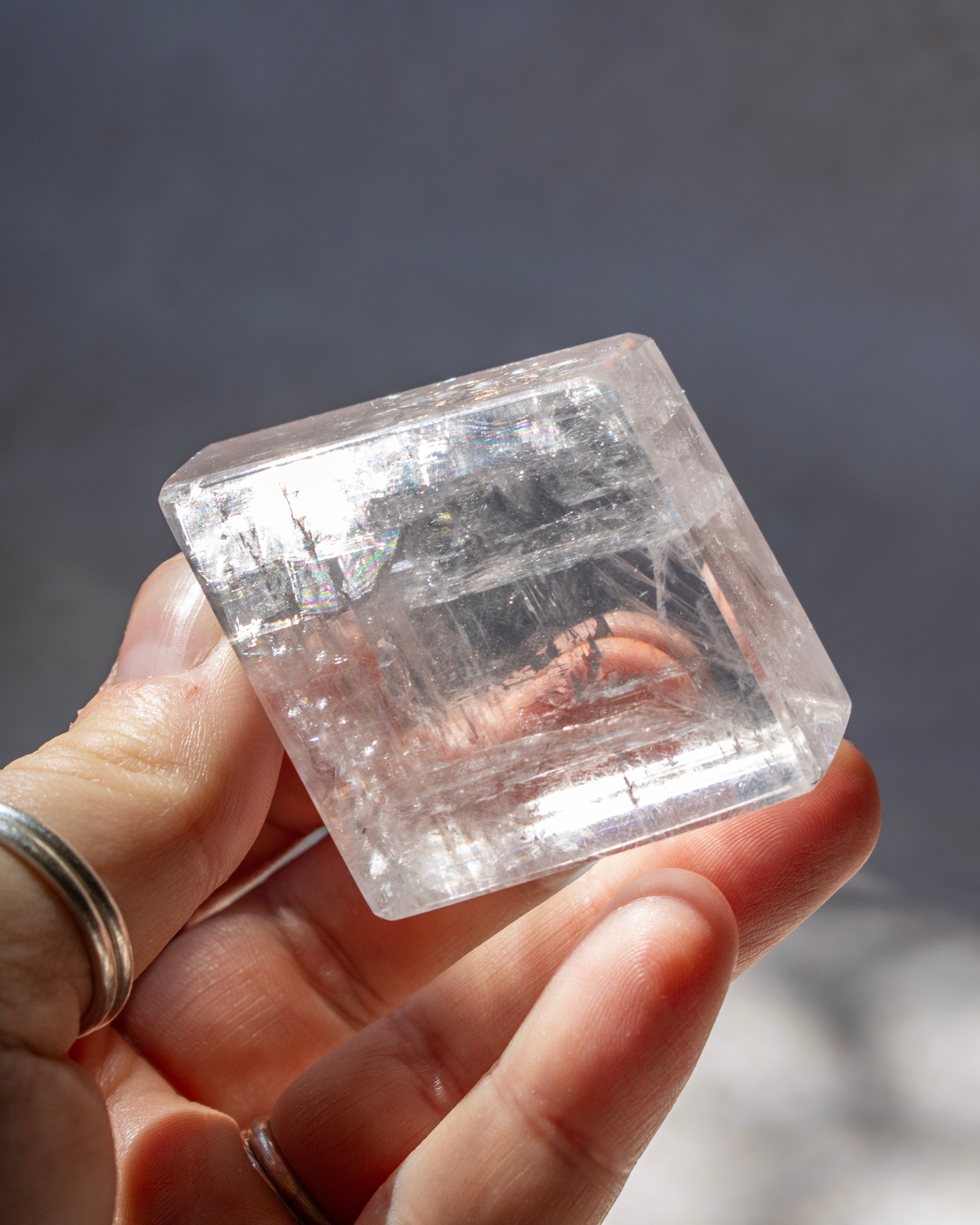 A hand holding a clear, faceted crystal cube with visible internal inclusions and natural striations, photographed against a soft gray background.
