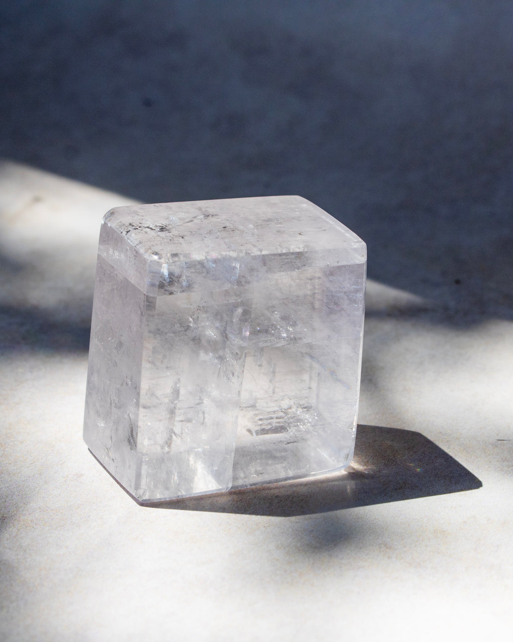 A clear, translucent cube-shaped crystal or stone block standing on a light surface with a dark shadow cast to the right, against a gray background.
