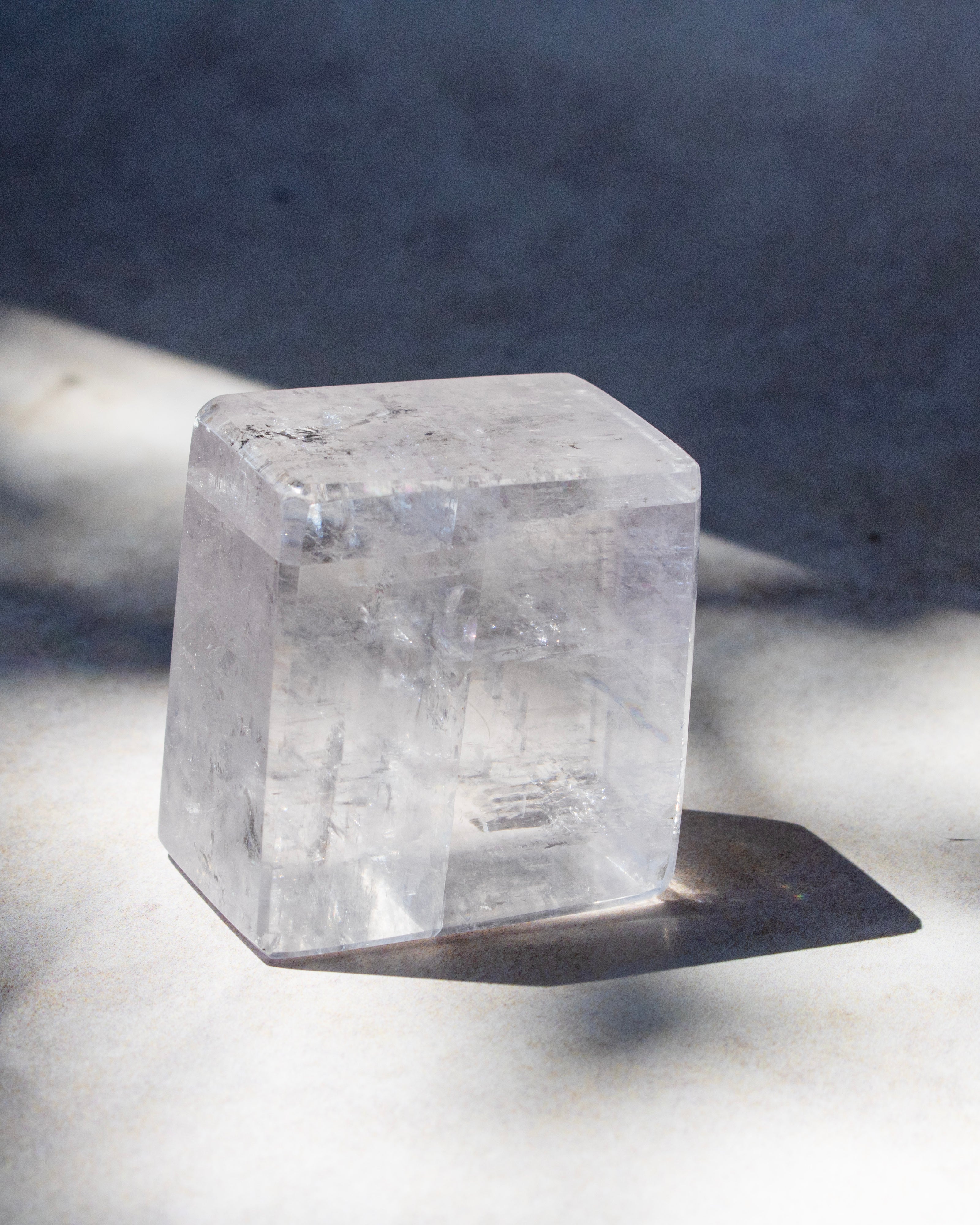 A clear, translucent cube-shaped crystal or stone block standing on a light surface with a dark shadow cast to the right, against a gray background.