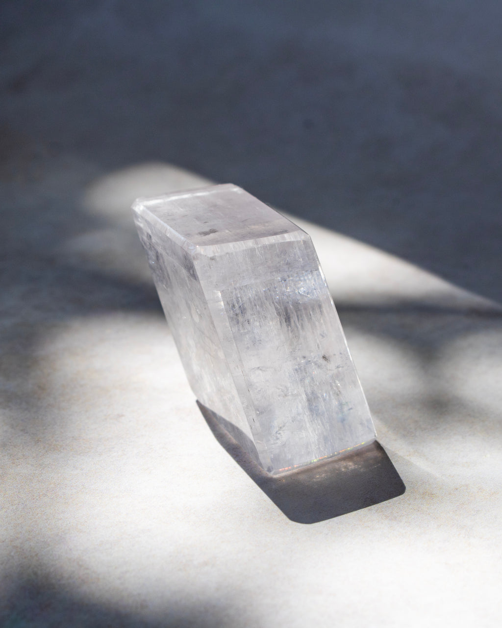 A clear rectangular crystal block or stone sample standing upright on a light surface, photographed in natural sunlight with strong shadows.