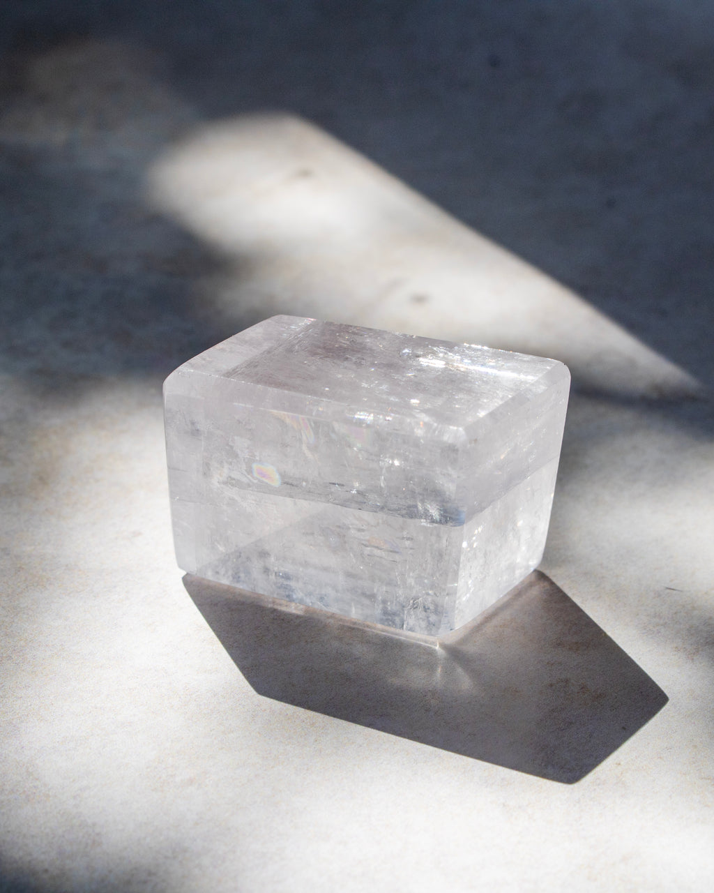 A clear rectangular crystal, photographed on a gray surface in dramatic natural sunlight with strong shadows.