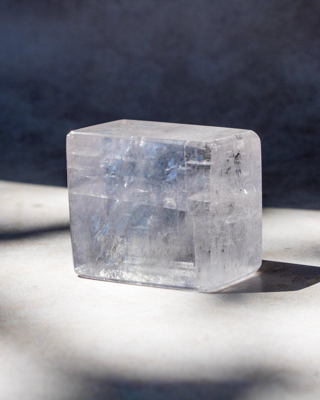 A clear, translucent square crystal or stone block with natural internal texture, photographed on a light surface with a dark blue-gray background and soft shadows.