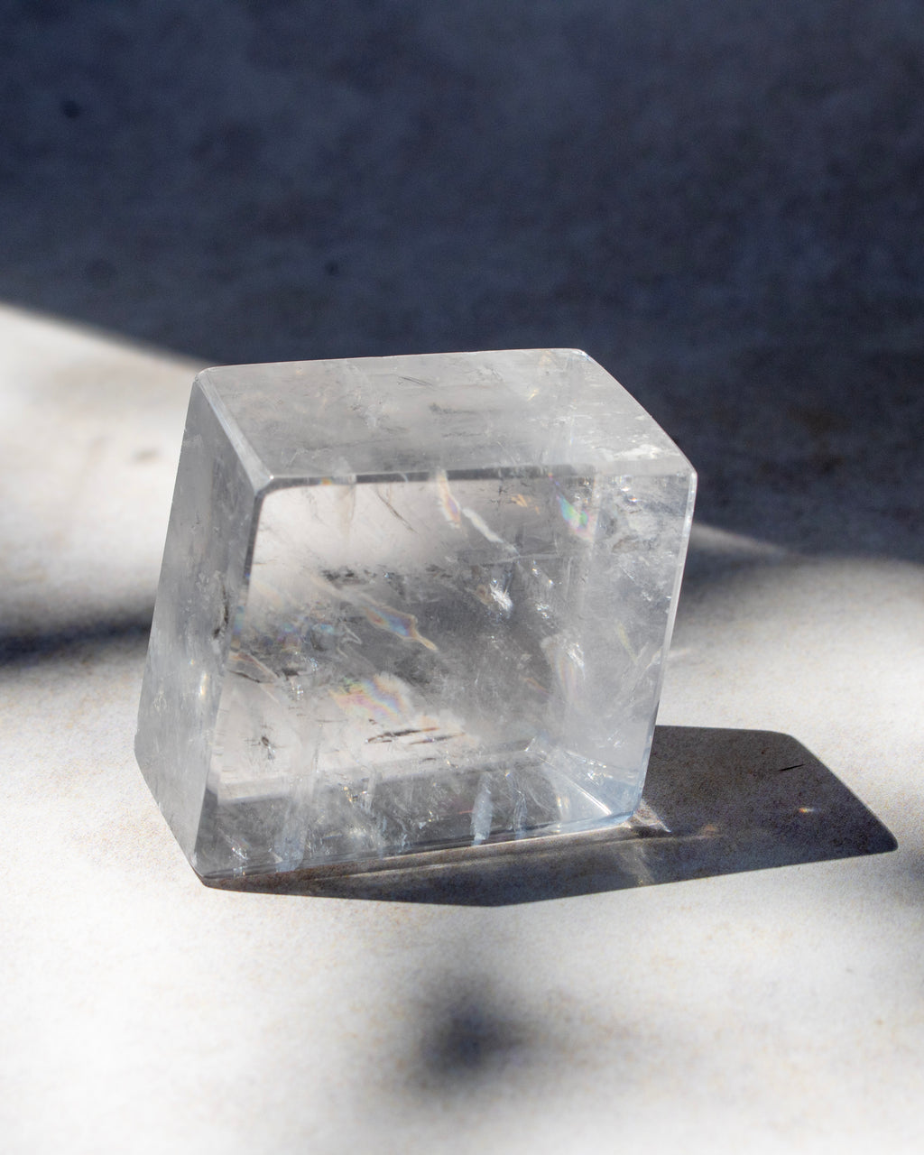 A clear, transparent cube-shaped crystal or glass block with natural internal inclusions, photographed on a light surface with a dark shadow cast to the right.