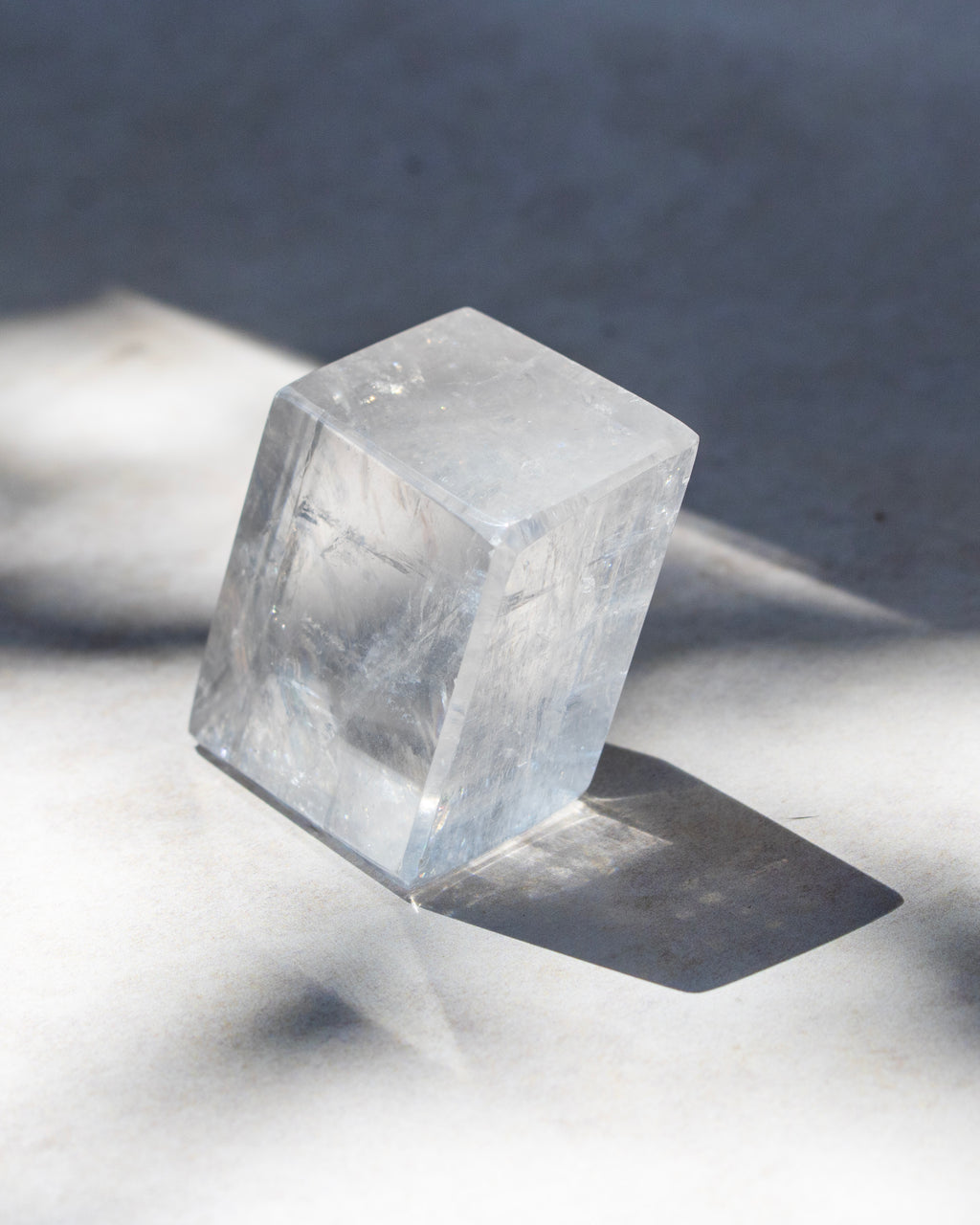 A clear, faceted crystal cube with natural internal inclusions, standing on a light gray surface under direct sunlight that casts a sharp shadow.