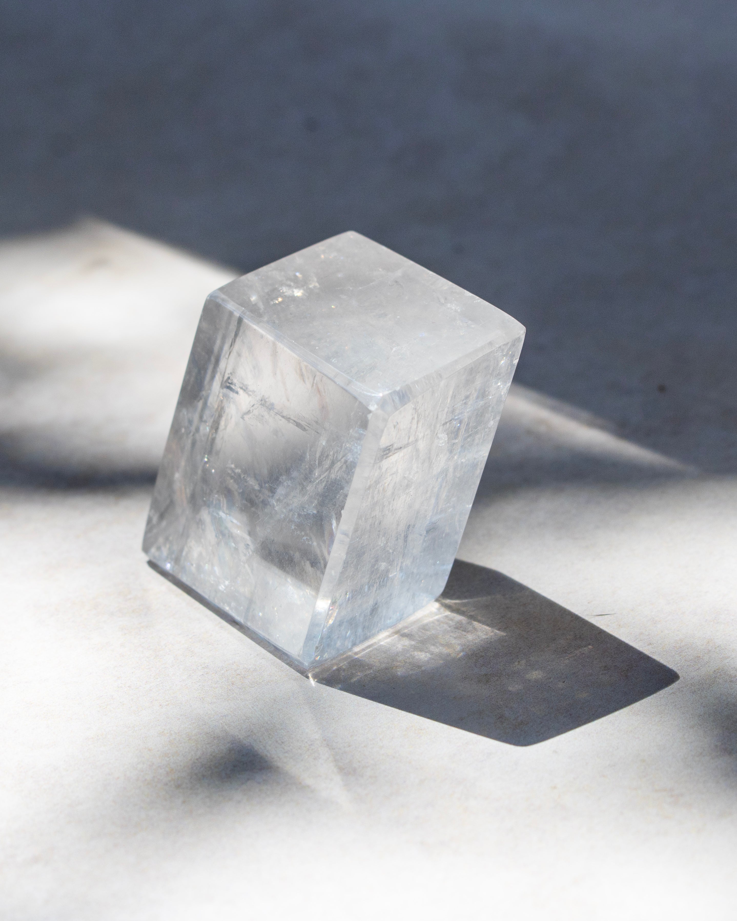 A clear, faceted crystal cube with natural internal inclusions, standing on a light gray surface under direct sunlight that casts a sharp shadow.